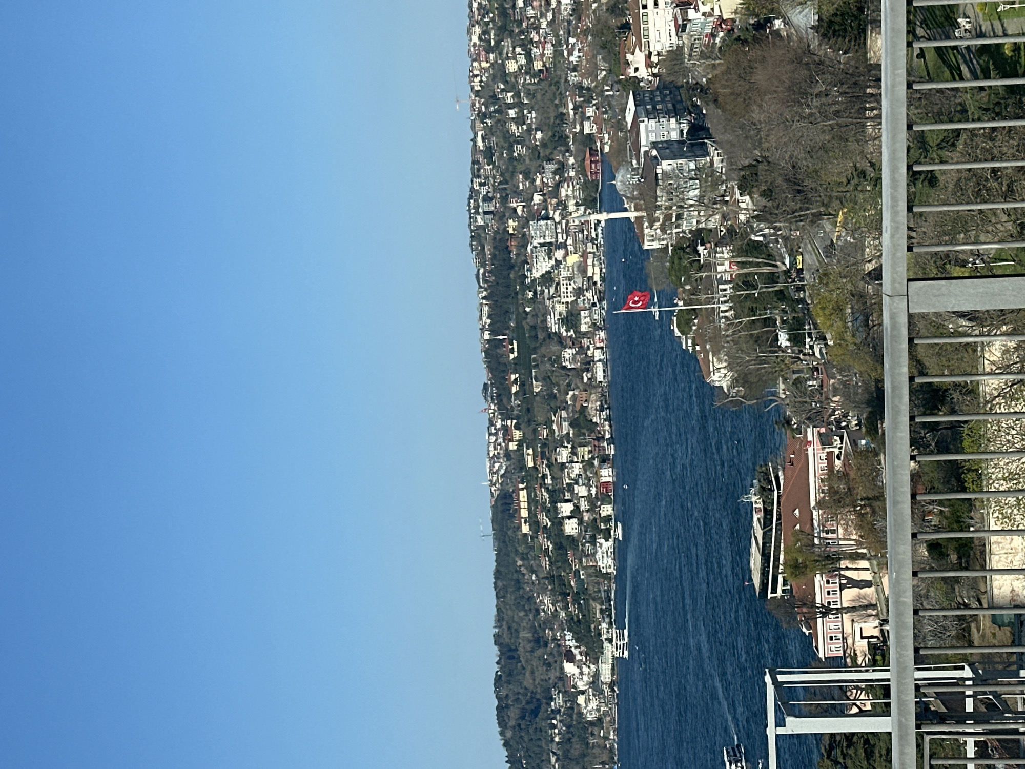 View of the Bosphorus strait in Istanbul, Turkey, taken from a high vantage point. The photograph shows a body of water, with a portion of the cityscape lining the coast on the opposite bank. Buildings are densely packed together and line the hills. A Turkish flag is visible near the center of the view. The sky is clear. A gray metal railing is visible along the right edge of the image. The image appears to be taken from a high location in the city of Üsküdar.