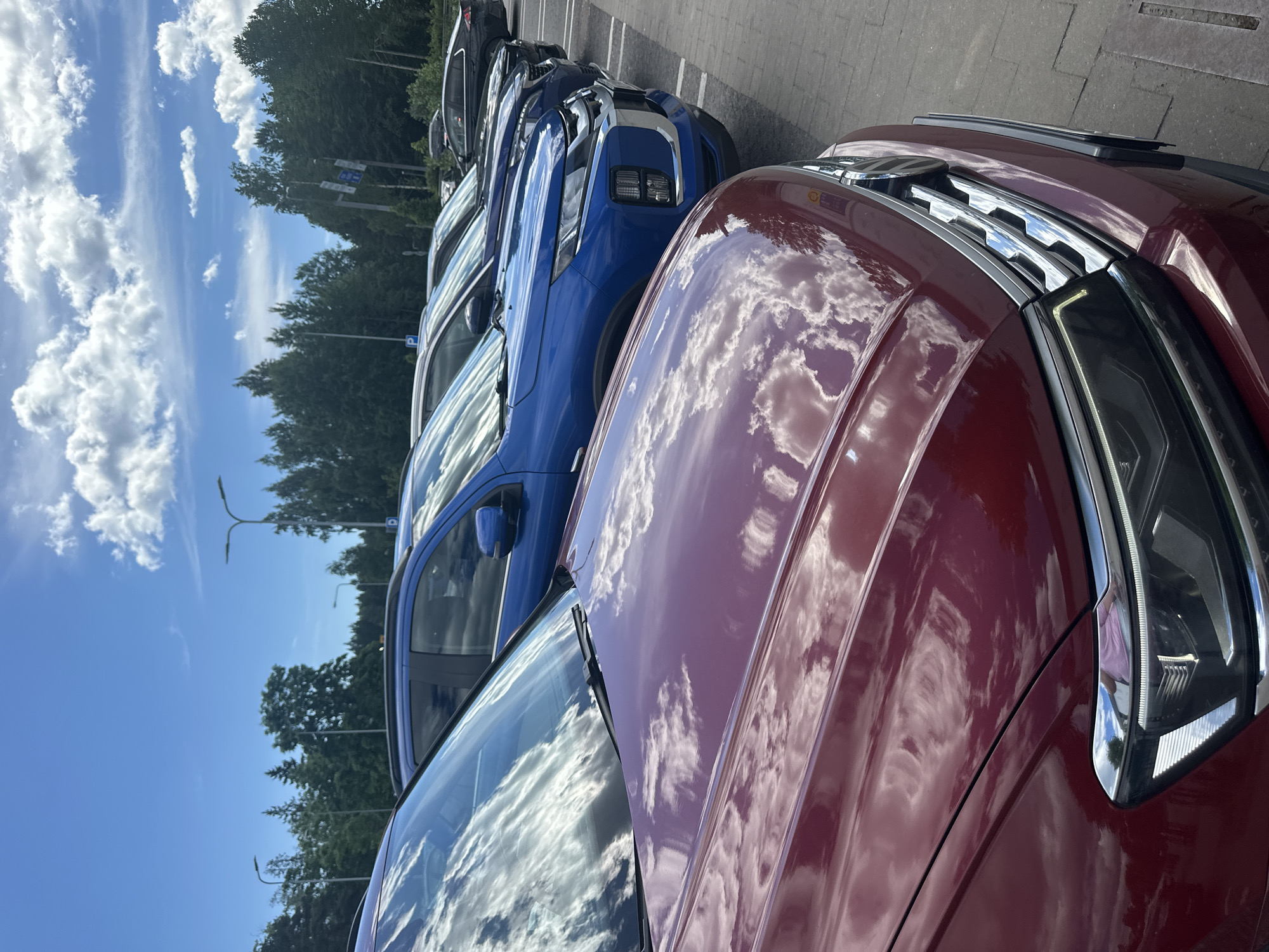 An outdoor, eye-level, medium shot captures a line of parked cars under a bright, cloudy sky. The closest car, a dark red SUV, is positioned in the foreground, reflecting the sky and clouds on its hood and windshield. Behind it, a line of other SUVs, including a bright blue one, are parked in a row. The background features a dense line of green trees and tall streetlights. The cars are parked on an asphalt surface. The overall scene is a typical parking lot setting.