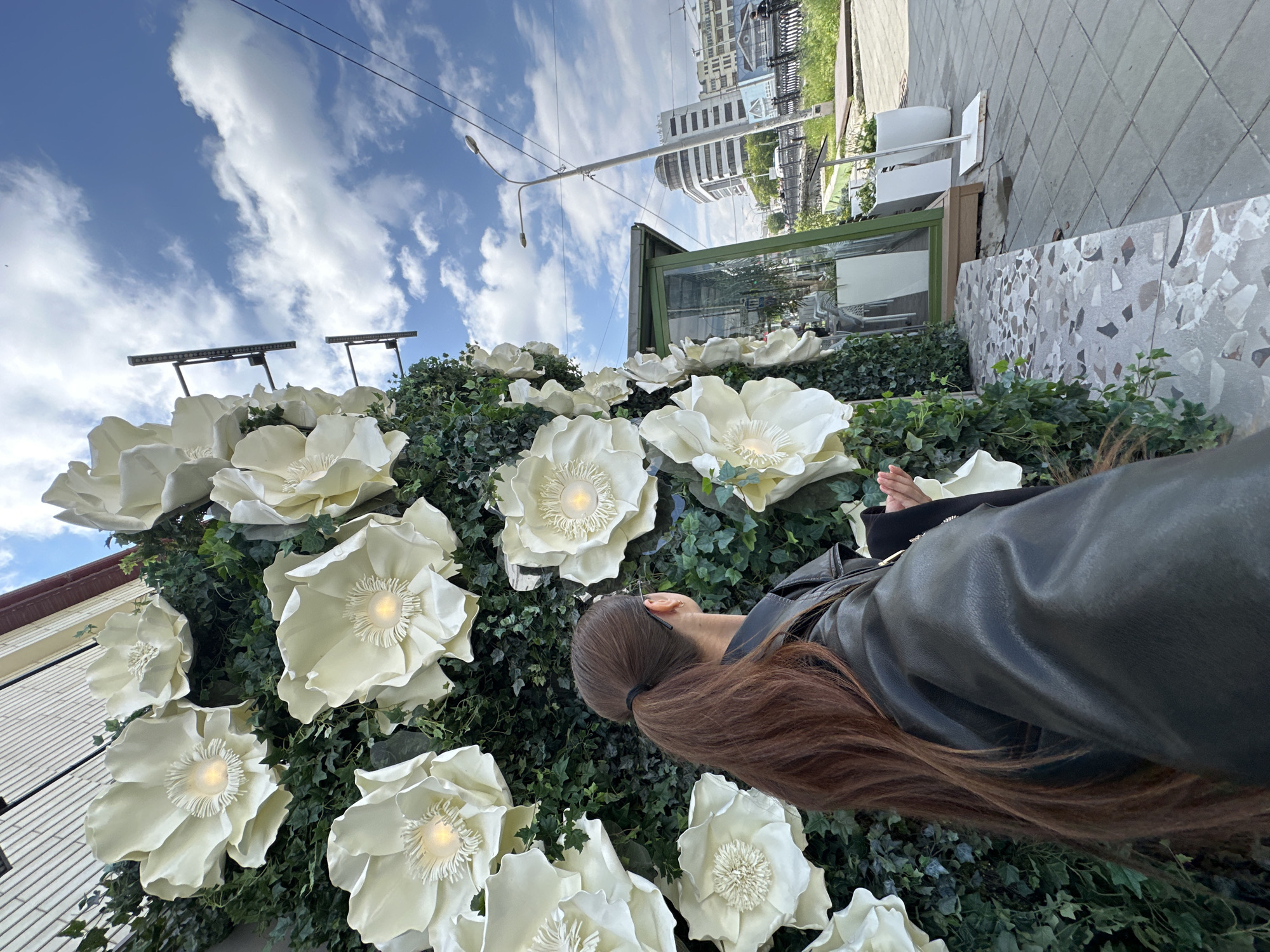 A woman with long, brown hair in a ponytail, wearing a black leather jacket, stands with her back to the camera, looking at a large, artificial flower display. The flowers are white and oversized, with illuminated centers, attached to a green wall of ivy. To the right of the display, a glass-enclosed structure, possibly a cafe, is visible, along with streetlights and buildings in the background. The scene is outdoors on a sunny day with a cloudy sky, on a sidewalk with a patterned tile border. The image was captured on June 9, 2024, at the Flower Cafe "Aivazovsky" in Yekaterinburg, Russia.
