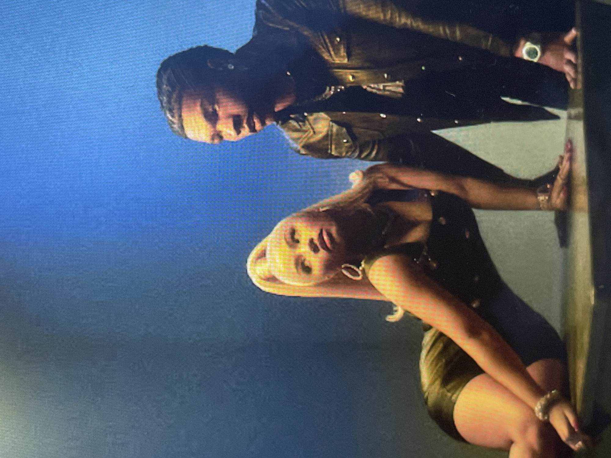 The image captures two individuals in a stylized pose within a dimly lit indoor environment, against a plain blue background that subtly shifts in tone. Although the image is presented with a 90-degree clockwise rotation, the scene, when viewed upright, portrays a man standing behind a bending woman.
The man, positioned towards the back, has dark hair and is wearing a dark, possibly leather, jacket or shirt adorned with metallic buttons. A prominent watch with a light-colored face is visible on his left wrist. He is looking downwards, his gaze directed towards the woman.
In the foreground, the woman has long, blonde hair and is wearing a dark, form-fitting outfit that appears to be a short dress or a combination of a top and skirt, possibly made of a reflective material. She is adorned with hoop earrings and bracelets on her wrists. Her body is bent forward, and her hands are resting on a dark surface or wall to her right. She is looking upwards with an intense, posed expression.
The overall activity suggests a choreographed moment, likely part of a photoshoot, a music video, or a performance. The lighting is dim, contributing to a dramatic and moody atmosphere. There are no specific architectural or geographical cues visible in the image itself, but the provided context indicates this scene could be taking place in Sofia, Bulgaria. No text is visible in the media file.