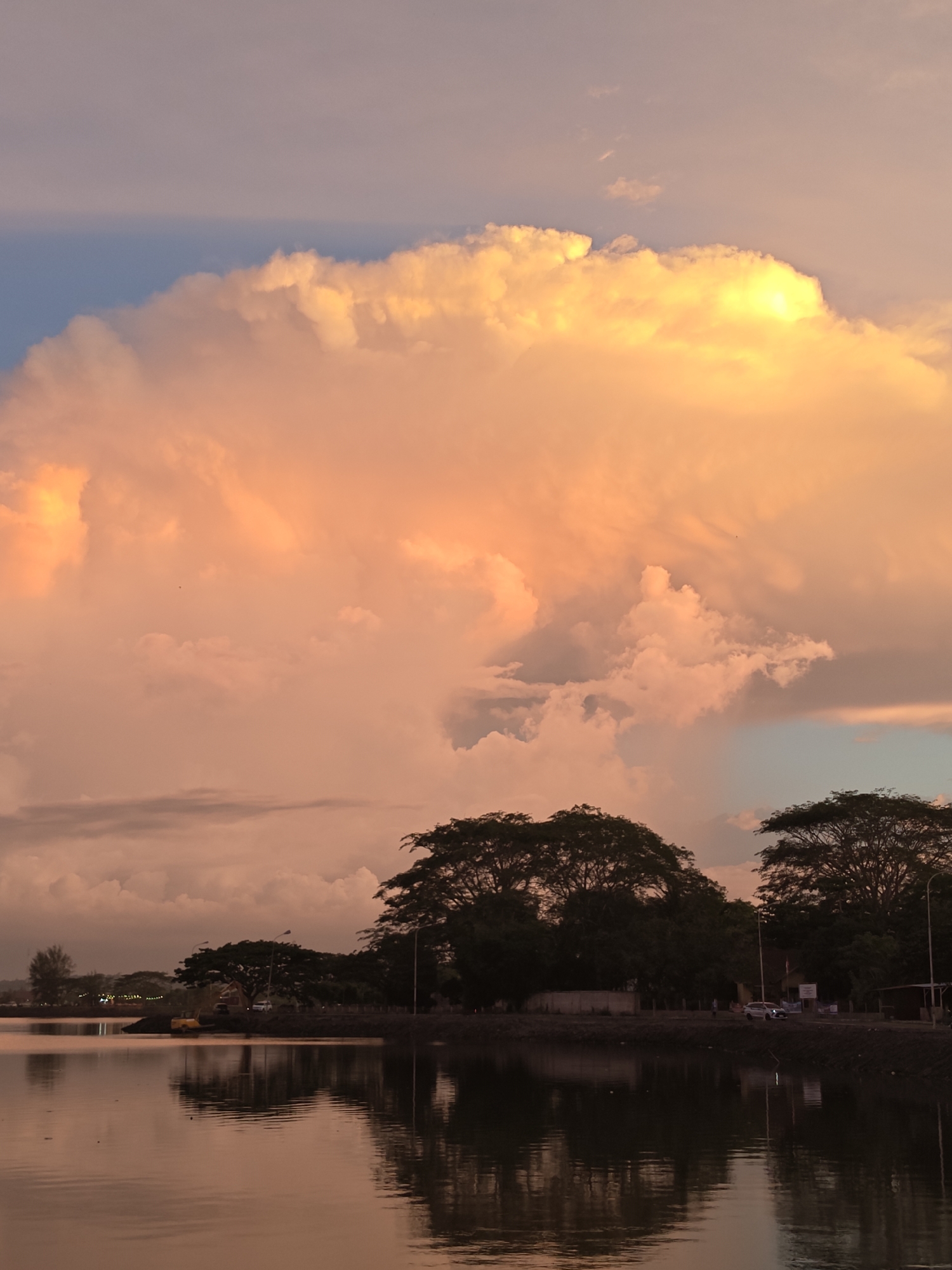 This is a vertical, medium-shot photograph of a serene waterscape at sunset, with Lhokseumawe, Indonesia, as the context. The dominant subject is a vast expanse of sky filled with dramatic, voluminous clouds. These clouds are illuminated by the setting sun, casting a warm, ethereal glow of orange, peach, and yellow hues across their surfaces, creating a breathtaking spectacle. The upper portion of the sky transitions from a soft blue to a hazy pastel, with a few wispy clouds scattered throughout.
Below the magnificent sky, a calm body of water occupies the lower half of the frame, mirroring the colors and shapes of the clouds and sky with gentle reflections. The water appears dark and still, creating a tranquil contrast to the vibrant sky. Along the far bank of the water, a line of silhouetted trees with full, dark canopies stands out against the lighter sky. The foliage of these trees is dense and lush.
Further back, behind the trees, a subtle hint of human activity is visible. Streetlights can be seen, and there are indications of a road or pathway. A few vehicles, including what appears to be a white car, are parked or driving along the bank, suggesting a roadside or park area. There are also faint lights in the distance, possibly from buildings or another settlement. One small, yellow object, possibly construction equipment, is partially visible near the water's edge.
The overall mood of the image is peaceful and awe-inspiring, evoking a sense of wonder at the natural beauty of a sunset. The time of day is clearly evening, as indicated by the sunset colors and the presence of artificial lights. The weather appears calm, with the still water suggesting little to no wind. The presence of the distinct trees and the overall landscape point towards a tropical or subtropical setting. No legible text is visible in the image.
