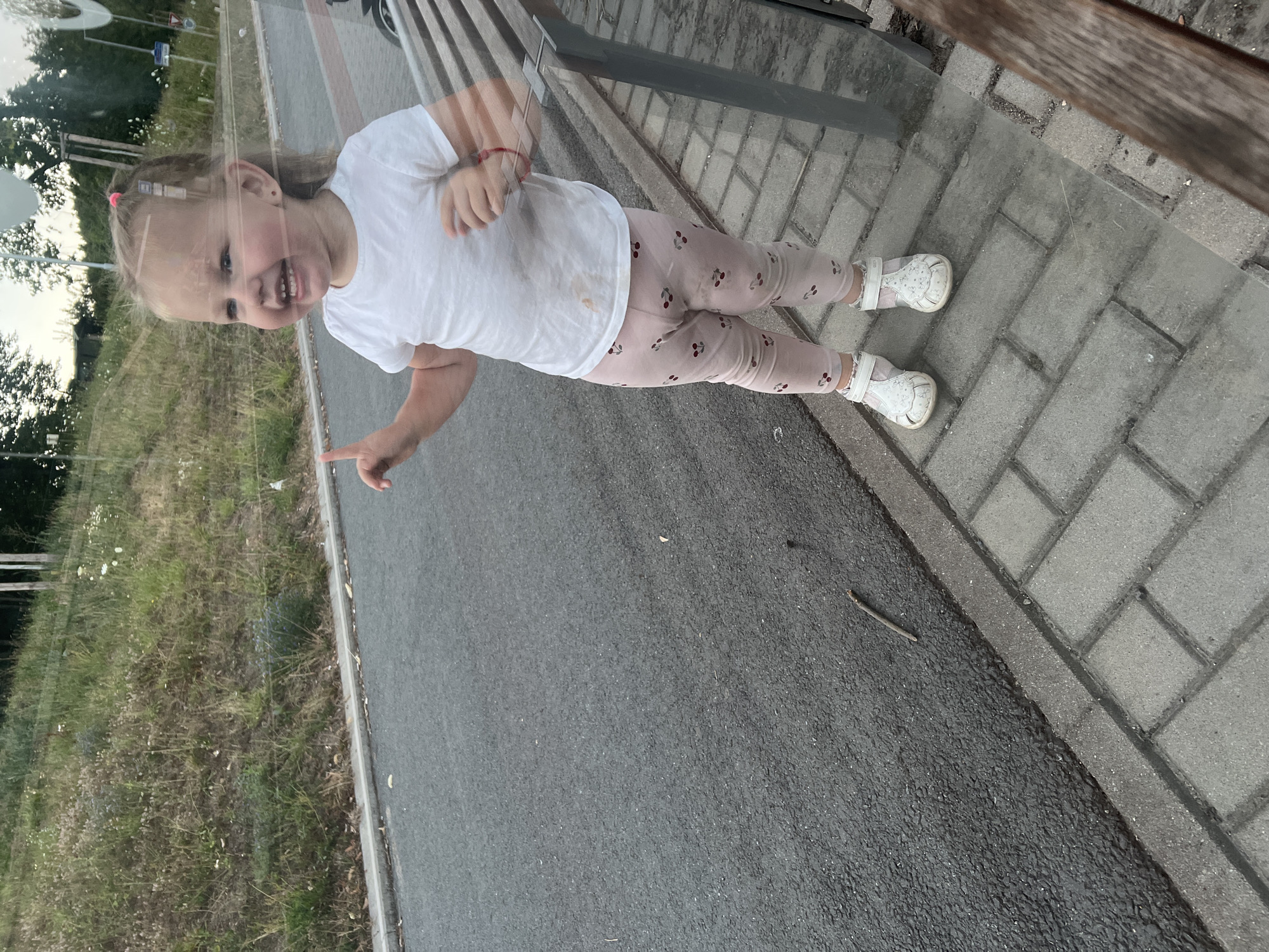 A young girl with blonde hair in a pink hair tie stands on a paved walkway next to an asphalt road, looking to her left and smiling with her mouth open. She is wearing a white t-shirt with a stain on the front, pink leggings with a cherry pattern, and white sandals. Her left index finger is raised. Behind her, a grassy slope is visible with some trees. A wooden bench is partially visible to her right, and a glass structure appears to be in front of her, creating reflections in the image. The scene is likely a street or park in Pilsen, Czechia, given the context.
