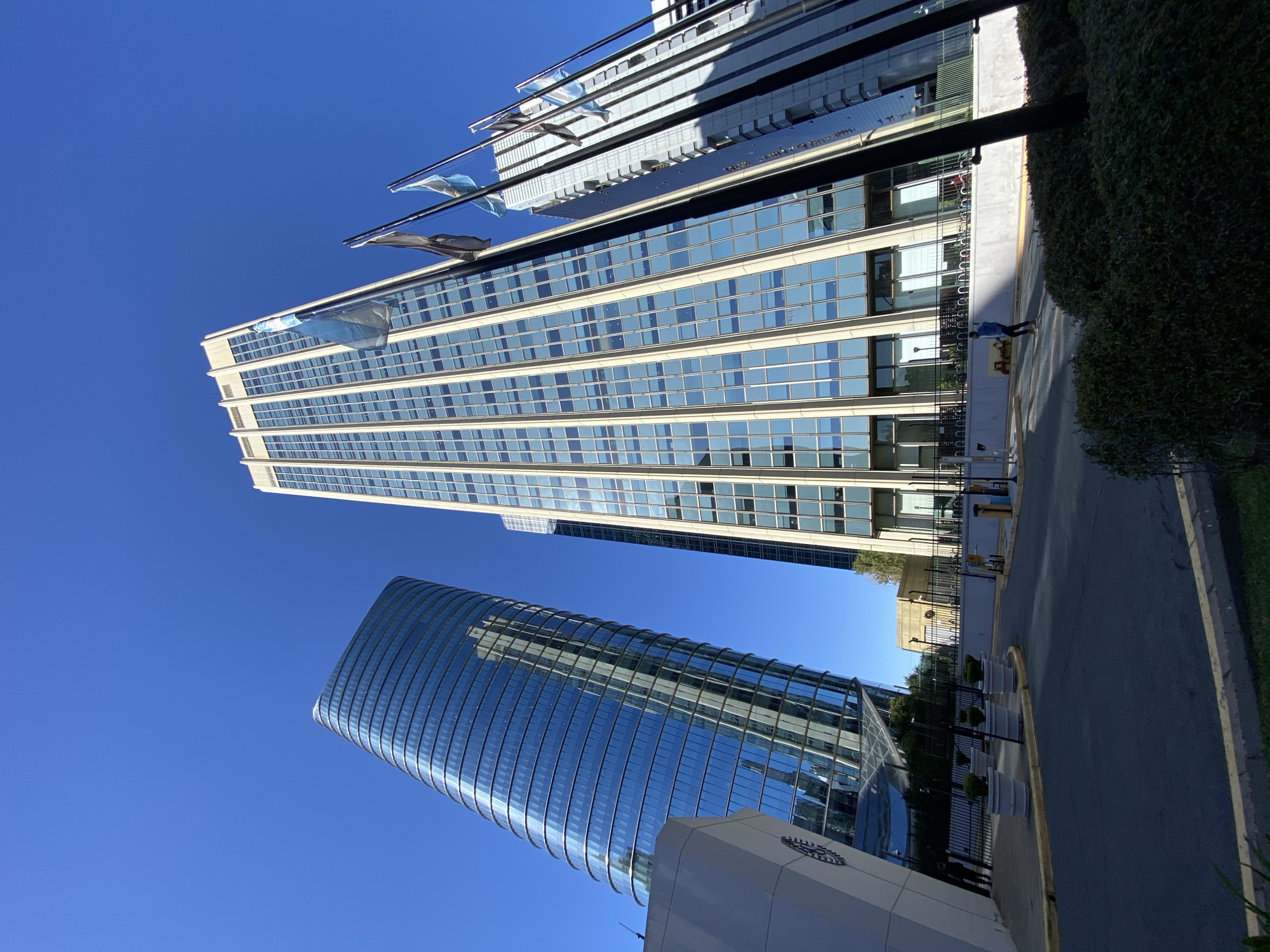 The image presents an urban landscape in Buenos Aires, Argentina, dominated by two modern skyscrapers under a clear, bright blue sky.
On the left, a tall, slender rectangular building features numerous horizontal rows of reflective windows that mirror the sky. Several light blue and white flags, likely representing Argentina, are attached to poles extending from the upper part of this building's facade.
To the right, a distinctively curved or cylindrical skyscraper, entirely clad in reflective glass, rises towards the sky, its surface also mirroring the surroundings. A white base with an unidentifiable circular logo is visible at the bottom of this building.
In the foreground, running along the base of the rectangular building, is a sidewalk and a road. An individual, appearing to be an adult male in a blue shirt, is walking on the sidewalk. A well-maintained green hedge or bush line the edge of the sidewalk, separating it from the street. Further down the street, several white cylindrical planters containing small green topiaries are placed alongside a black fence.
The bright sunlight casts distinct shadows, indicating it is daytime, likely late morning or early afternoon, with clear weather. The scene portrays a modern, active city environment. No discernible text is clearly legible in the image.