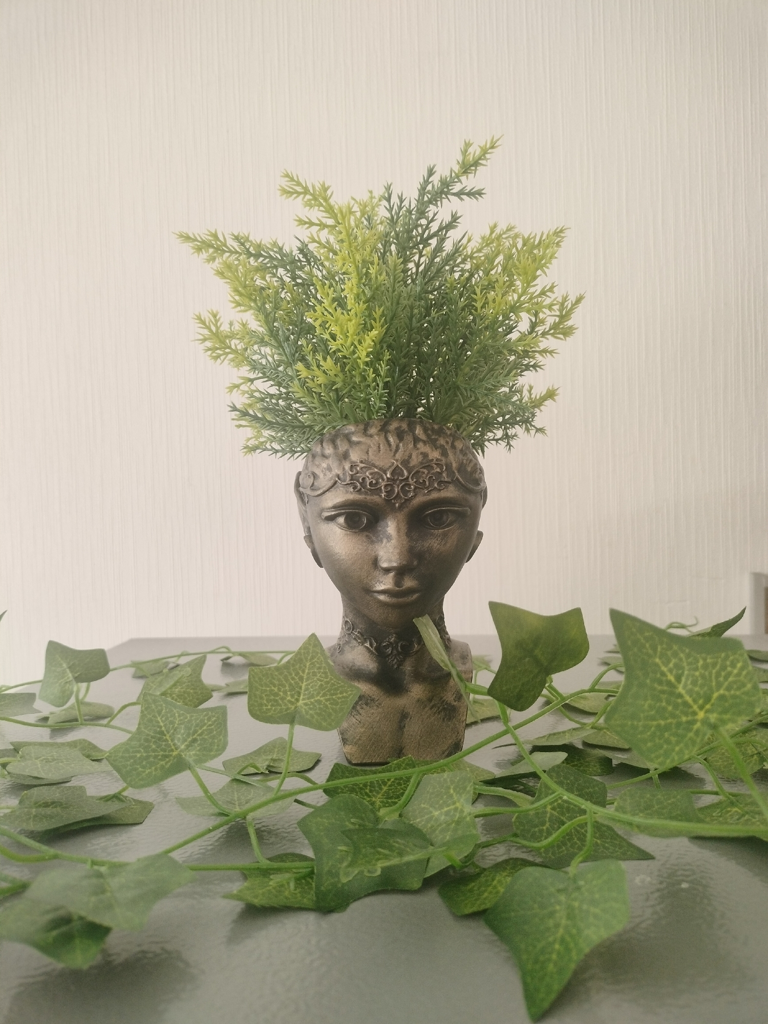 An antique-style bust of a woman with a plant growing from her head sits on a flat surface. The bust is made of a metallic, bronze-colored material and has intricate detailing on the forehead and neck. The plant is a vibrant green with bushy, fern-like leaves. Several artificial ivy vines with heart-shaped leaves are scattered around the bust on the surface. The background is a plain, textured wall. This image was taken indoors, in the city of Lyubertsy, Russia.