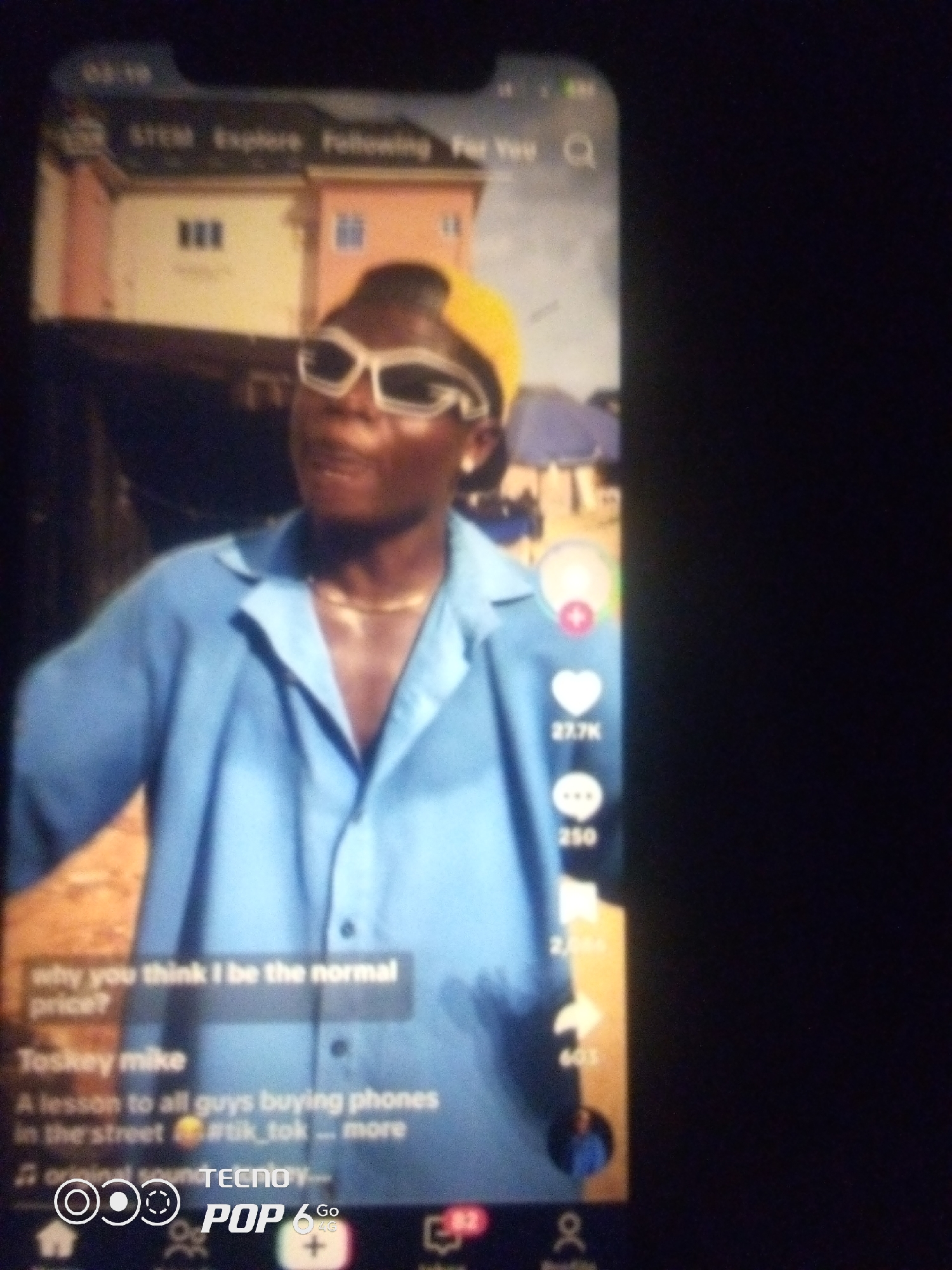 The image shows a screenshot of a TikTok video playing on a smartphone. The video features a Black male wearing a light blue collared shirt and a yellow durag. He is also wearing white-rimmed sunglasses and a gold chain necklace. The background of the video appears to be a house with a light pink and yellow facade, situated on stilts, suggesting a location with water. The text overlay on the video reads "why you think I be the normal price? Toskey mike A lesson to all guys buying phones in the street #tik_tok --- more." On the right side of the screen, there are social media interaction icons, including a heart with "27.7K" likes, a speech bubble with "250" comments, and a share icon with "603" shares. The bottom of the screenshot shows the TikTok interface with navigation icons and branding that includes "TECNO POP 6 Go". The image is blurry and taken at an angle, with a dark background suggesting the surrounding environment. The prompt mentions Howayi, Nigeria, but this information is not visually verifiable from the image itself.
