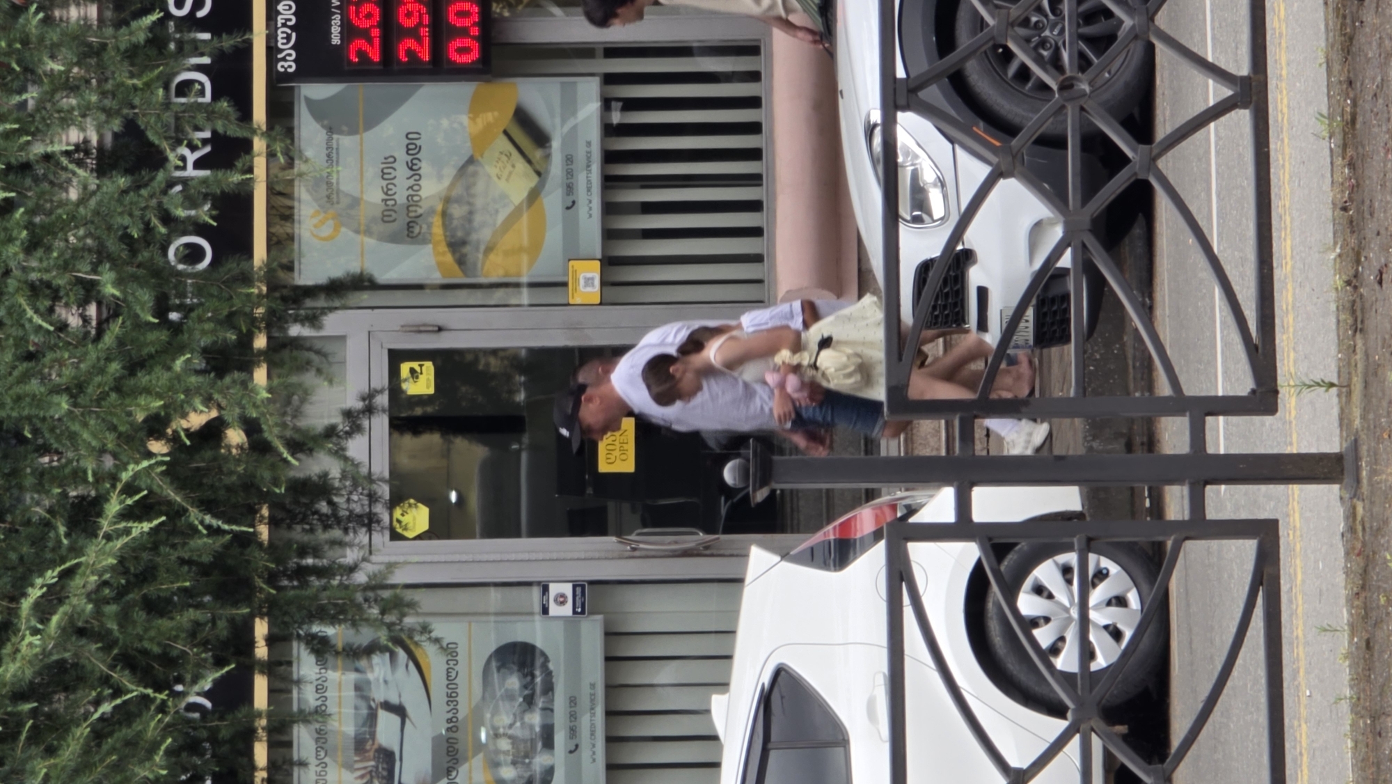 The image depicts a street scene in Batumi, Georgia, with a building featuring a storefront for "CREDITS." A man wearing a white t-shirt, blue shorts, and a black baseball cap is walking with a young girl in a light-colored dress. The girl is holding a pink object. They are walking down a short set of stairs. In the foreground, a black metal fence is visible, and behind it, parts of two white cars are parked on the street. The building's facade shows signage with Georgian text and a digital display showing currency exchange rates, with numbers like "2.6" and "2.9" illuminated. There are also signs indicating "OPEN" and security camera logos. Evergreen branches hang over the top of the building.