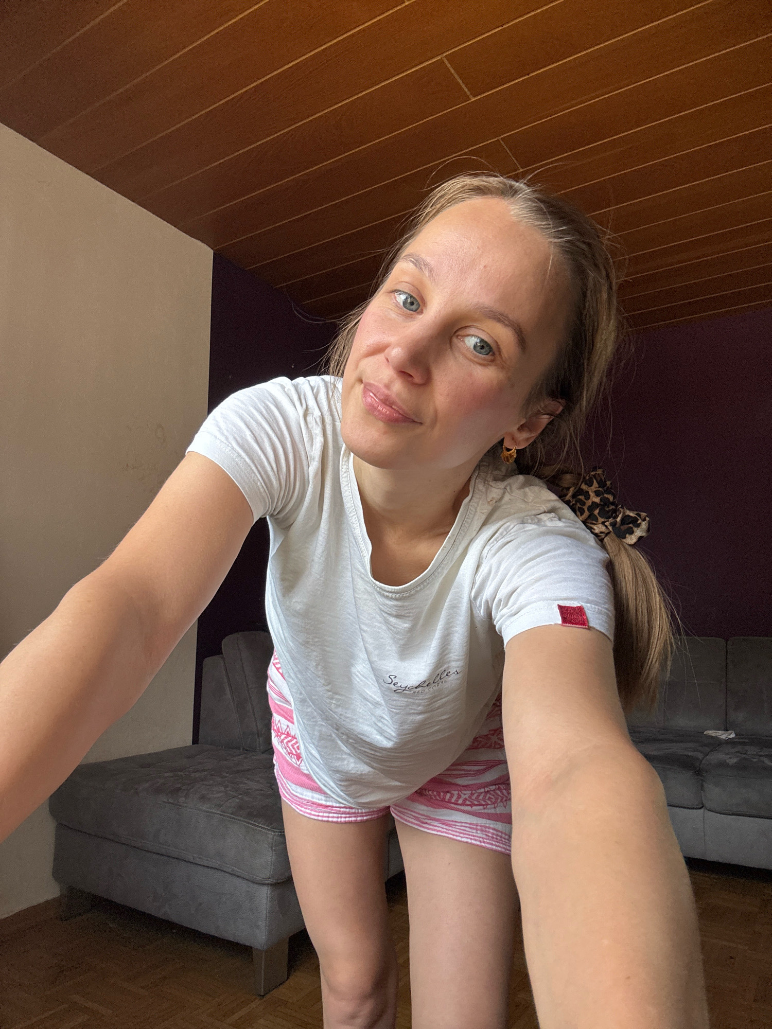 A young woman with blonde hair pulled back in a ponytail with a leopard print scrunchie is captured in a selfie. She is wearing a white t-shirt with "Seychelles" printed on it in cursive and pink and white striped shorts. She has a light tan and is smiling at the camera, with her left arm extended. Behind her, a grey couch is visible in what appears to be a living room. The room has a wooden slatted ceiling and a dark purple wall. The floor is made of hardwood. The image seems to be taken indoors during the daytime.