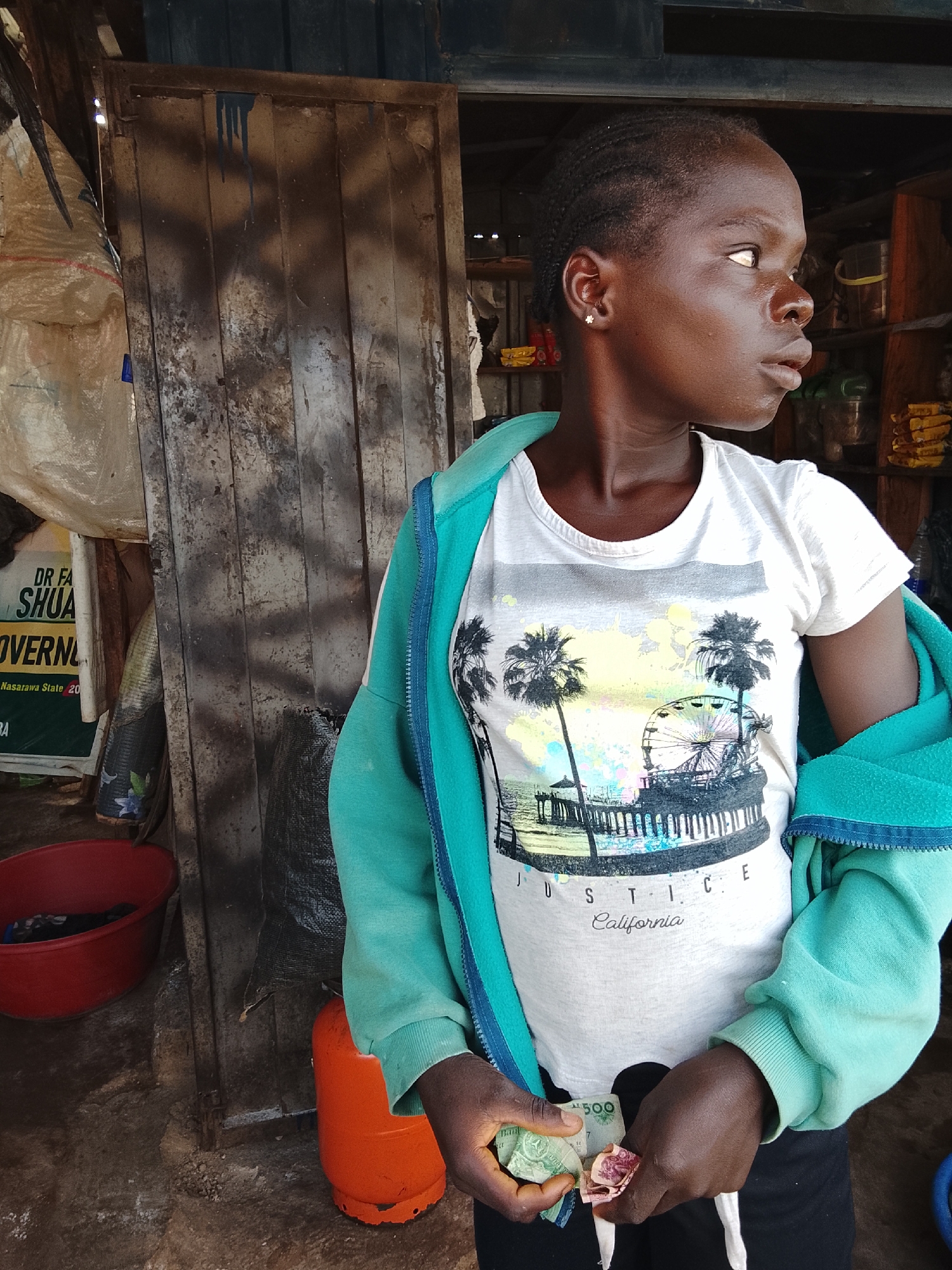 A young person stands in front of a weathered wooden door, possibly a shop entrance, in Akwanga, Nigeria. The person wears a white t-shirt with a graphic print featuring palm trees, a pier, and a Ferris wheel, along with the words "JUSTICE California". A light blue hooded jacket is draped over their shoulders. They are holding several crumpled banknotes in their hands, appearing to be counting or examining them.
The background is rustic and appears to be an outdoor market or a small storefront. To the left, a large white bag with red stitching is visible, along with a poster with visible text including "DR FA SHUA GOVERN" and "Nasarawa State 20". A bright red basin is also present. To the right of the person, shelves with various items are visible inside the shop. An orange gas cylinder stands on the ground near the person's feet.
The lighting suggests it could be daytime, with natural light illuminating the scene. The overall impression is that of a candid moment captured during a transaction or while the person is preparing to make a purchase. The person's gaze is directed off-camera to the right, with a neutral or perhaps slightly contemplative expression. The environment appears somewhat unkempt, consistent with many open-air markets in Nigeria.