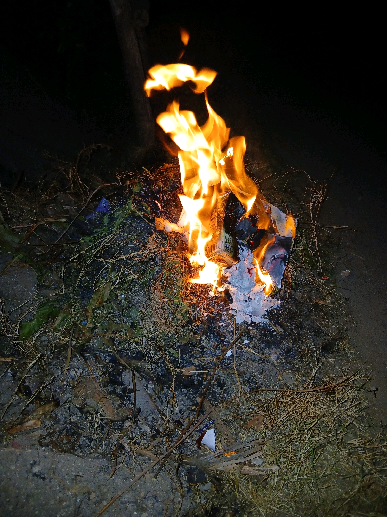 This is a nighttime, medium-angle, close-up shot of a small, contained fire burning on the ground. The fire is primarily composed of bright orange and yellow flames, with some white and grey ash visible at its base. The materials being burned appear to be dried vegetation, such as grass and twigs, along with some paper products, including a small cardboard box or container with some text visible. The ground surrounding the fire is a mixture of dirt, ash, and scattered organic debris. The background is dark, suggesting an outdoor environment at night. There are no people or animals visible in the image. The scene is consistent with an outdoor bonfire or the burning of refuse in a semi-urban or rural area. Without specific geographic markers, the location in Pangkalpinang, Indonesia, cannot be definitively confirmed from the image alone.