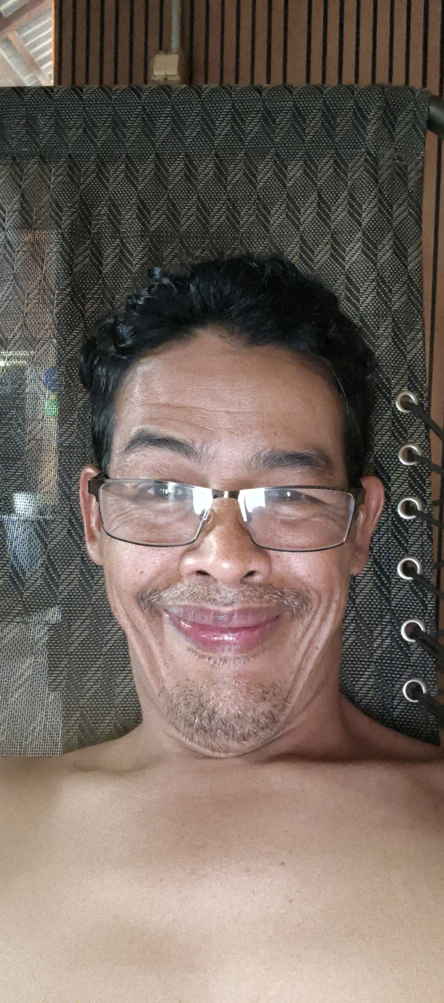 An individual with dark, curly hair and glasses smiles at the camera. The person is shirtless and appears to be outdoors, seated on a chair with a patterned fabric back. The background shows vertical wooden planks. The location is Muaro Jambi, Indonesia.