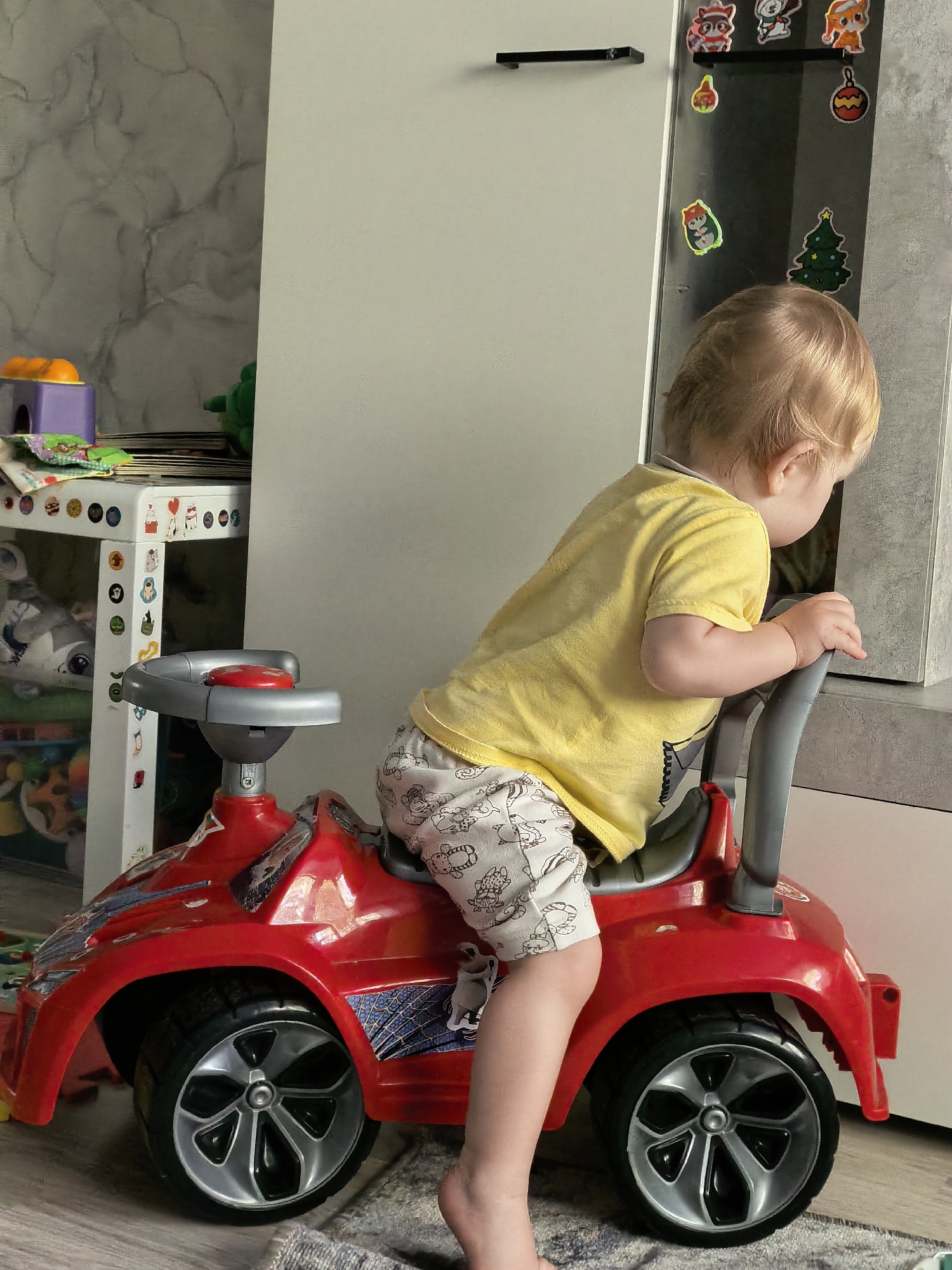 A young Caucasian child with blonde hair, wearing a yellow t-shirt and light-colored shorts with a cartoon pattern, sits on a red toy car. The child is facing away from the camera, looking towards a refrigerator adorned with various cartoon stickers, including a Christmas tree and animal characters. The toy car has silver accents and black wheels. The background includes a white cabinet, a grey, marbled wall, and a small white table with toys on it. The scene appears to be indoors, possibly in a residential setting. The image was taken in Sterlitamak, Russia.