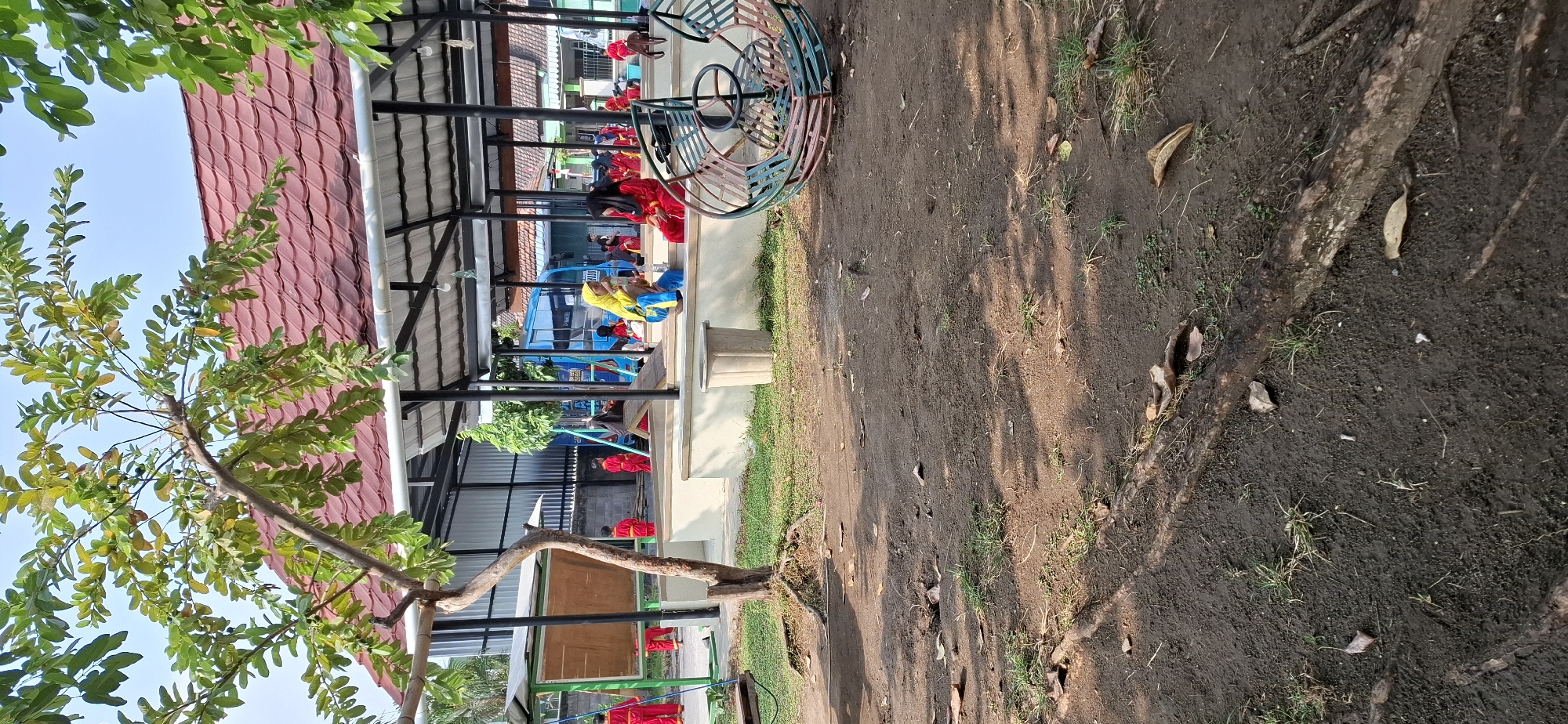 The image captures an outdoor scene at a playground or school in Polokarto, Indonesia. The foreground is dominated by a dirt ground with visible tree roots and fallen leaves, suggesting a natural or semi-maintained environment. In the mid-ground, there is a covered seating area with a red tiled roof supported by dark metal poles. Several children are present, some wearing red uniforms, and others in yellow and blue. They appear to be engaged in various activities, with some sitting and others standing. To the left, a tree with green leaves and visible fruit grows prominently. Behind the seating area, a blue bus is parked, and in the background, buildings with windows and roofs are visible. A circular playground structure, painted in muted colors of green, brown, and pink, is positioned on the right side of the mid-ground. The sky is a clear blue, indicating fair weather.