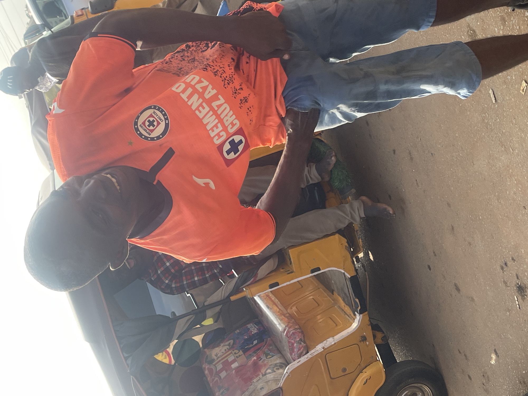 A group of people are gathered around a yellow auto-rickshaw, commonly known as a 'Keke Napep', in what appears to be an outdoor market or roadside setting in Galadimawa Village, Nigeria. The focal point is a man wearing an orange t-shirt with a graphic print and blue denim shorts, who is partially obscured. Another individual is visible in a red and black checkered shirt. The Keke Napep is loaded with various items, including what looks like fabric rolls or packages. The ground is a rough, sandy surface, indicative of a rural or semi-urban environment. Sunlight suggests it is daytime. The atmosphere seems to be one of transaction or preparation for travel, with people interacting with the vehicle and its contents. The man in the orange shirt has a visible logo on his shirt that reads "CEMENTO CRUZ AZUL".