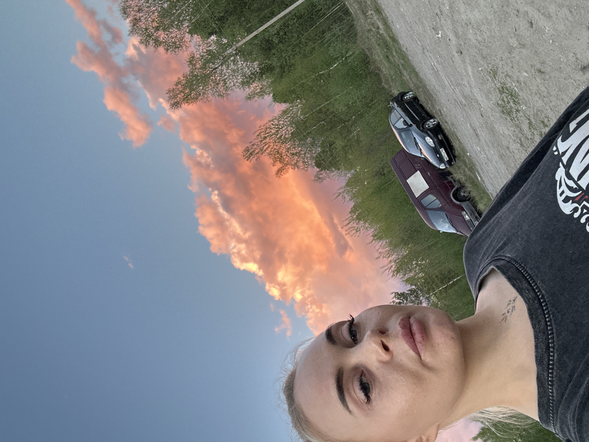 A blonde woman is captured in a selfie, with her face occupying the lower right portion of the frame, looking towards the camera. She has defined eyebrows and subtle makeup, and a small, intricate tattoo resembling a plant or branch design is visible on the left side of her neck. She wears a dark grey or charcoal t-shirt with a white, stylized graphic on the shoulder/chest area, though the text is unreadable.
Behind her, a striking sunset or sunrise sky dominates the background, featuring vibrant orange and pink clouds against a clear blue. To the mid-ground and left, lush green trees line what appears to be a dirt or gravel road. Two cars are parked on this unpaved surface: a dark-colored sedan or hatchback positioned further back, and a maroon sedan closer to the foreground. The overall setting suggests an outdoor, possibly semi-rural or suburban location, consistent with Vysokiy, Russia. The time of day is clearly twilight, marked by the dramatic, colorful clouds.