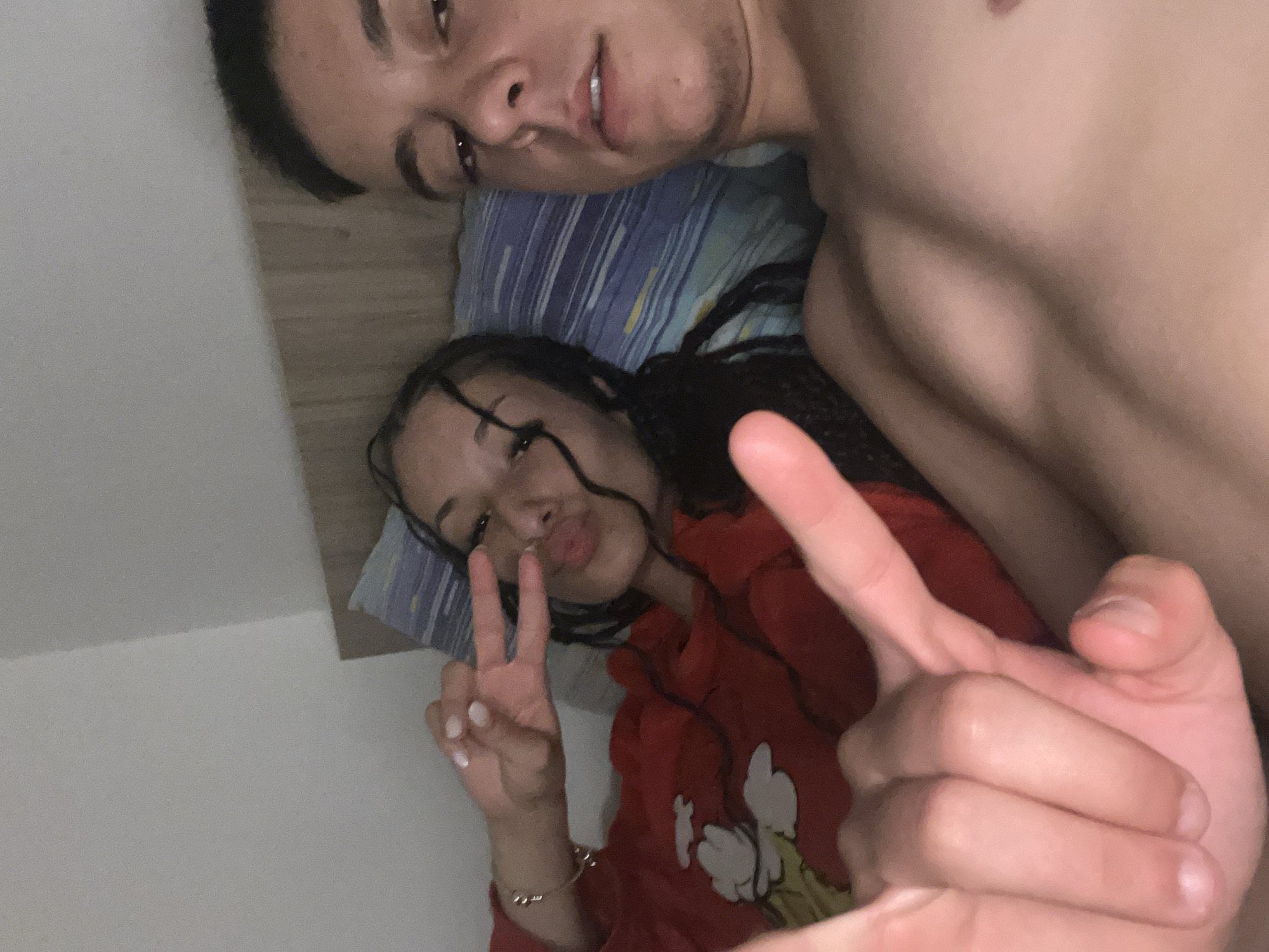 A close-up shot depicts two individuals lying in bed. One person, with dark hair, is positioned at the top of the frame, their upper body visible. The other person, with braided hair, is positioned below, wearing a red shirt, and making a peace sign with their fingers. They are lying on a pillow with a blue and white striped pattern. The location appears to be indoors, within a bedroom setting, but the specific city is not discernible from the image.