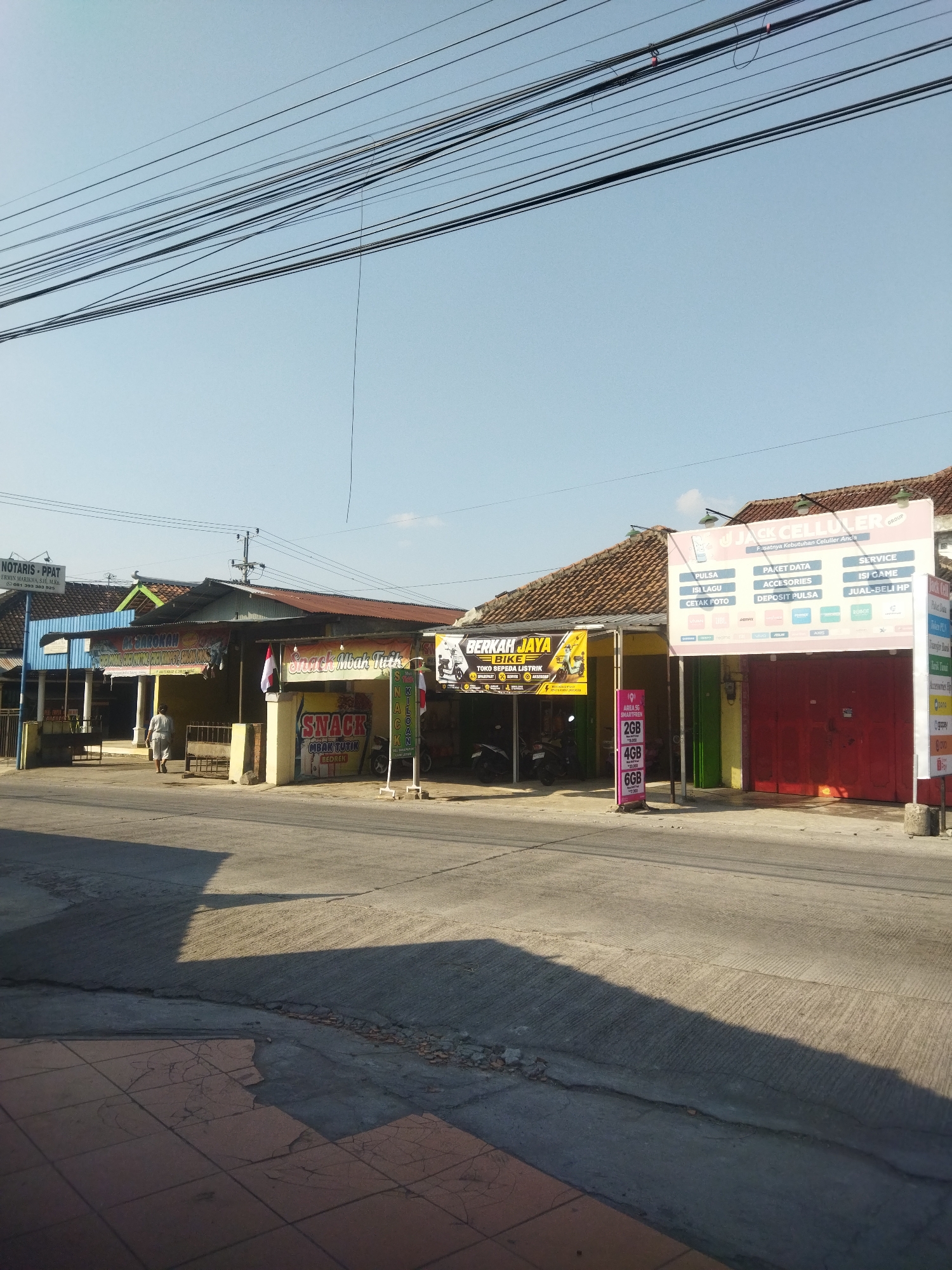 This image shows a street scene in Juwiring, Indonesia, with several small shops lining the right side of the road. The shops appear to be a mix of businesses, including a notary office, a food establishment, and a cellphone store. Overhead, a dense network of electrical wires crosses the frame. The road itself is paved and shows signs of wear, with some cracks and debris. Sunlight casts long shadows across the road, indicating it is likely late afternoon.
Specifically, on the left, there is a building with a blue awning and a sign that reads "NOTARIS - PPAT". Further down the street, a person is walking away from the camera towards a building with a yellow facade and a banner that reads "AL-HAROKAH". Next to this, a shop with a red front and a sign for "JACK CELLULER GROUP" is visible, advertising phone services and accessories. To the left of the "JACK CELLULER GROUP" shop, there is a display stand with various data package sizes (2GB, 4GB, 6GB) and their prices. A motorcycle is parked near the entrance of the "BERKAH JAYA BIKE" shop. Another sign reads "SNACK Mba Ttuk" with additional text that appears to be related to food items. A national flag of Indonesia is displayed between the "AL-HAROKAH" and "SNACK Mba Ttuk" shops. The pavement in the foreground consists of red tiles, some of which are cracked.