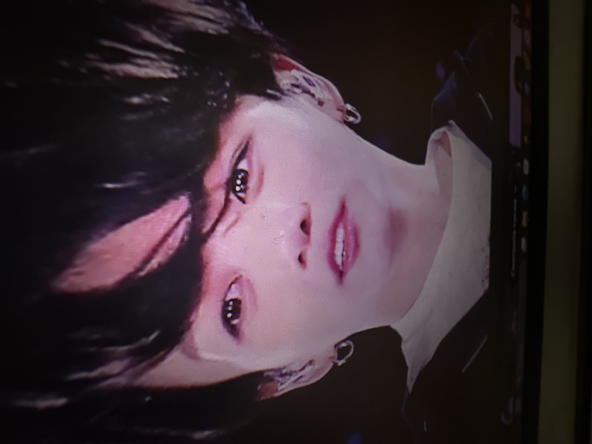 The media file presents a rotated close-up portrait of a person's face, appearing to be a photograph taken of a digital screen. The subject has dark hair, fair skin, dark eyes, and light pink lips slightly parted, revealing teeth. Their hair is swept across the forehead. Multiple earrings are visible; the left ear features several small studs or hoops, while the right ear has a single silver hoop earring. A light-colored collar or fabric is visible on the right side of the person's neck. The background is dark and out of focus, emphasizing the subject. The person's expression is direct and intense. Visible pixelation and the edge of a computer monitor with faint vertical interface elements are present on the right side of the image, indicating it is a screen capture or a photo of a display. The setting appears to be an indoor, controlled environment, likely for a photoshoot or video. No discernible text is visible.