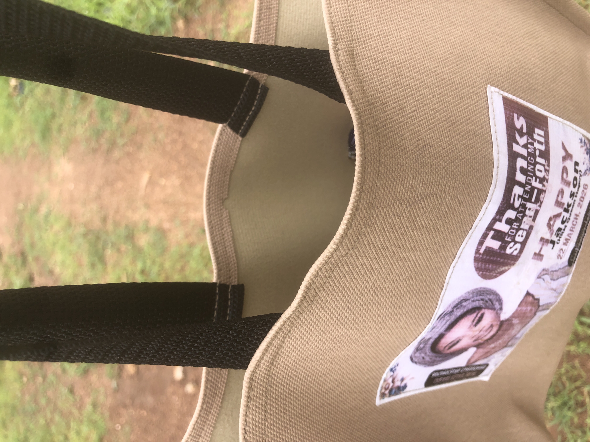 This media file is a close-up, slightly angled shot of the inside of a tan canvas tote bag. The bag has two black fabric straps. A sticker is attached to the exterior of the bag. The background is blurred and appears to be outdoors, with green grass and brown earth visible.
The sticker on the bag has text and an image. The text reads "Thanks for attending my Birth Happi", followed by "22 MARCH, 2020". A portion of the image on the sticker shows a person, likely a woman, wearing a hat and a patterned garment. The person's face is visible, and they appear to be smiling. The overall impression of the sticker is that it is a party favor or a thank-you note for an event. The date suggests the event took place in March 2020. The location context is Jalingo, Nigeria. The lighting suggests it is daytime, and the outdoor setting with grass implies fair weather.