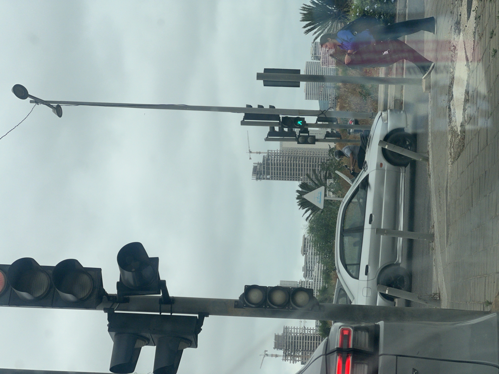 The image depicts an overcast daytime street scene in Casablanca, Morocco, captured from inside a vehicle, as indicated by visible reflections. The perspective looks towards an intersection and an urban backdrop.
In the immediate foreground, the rear red taillight of a dark vehicle, presumably the one from which the photo was taken, is visible on the right. Above this, a large pole spans the width of the road, supporting various traffic lights and streetlights. On the left side of the pole, a traffic light cluster displays a red signal. To the right, another set of traffic lights appears unlit, while a pedestrian signal on a separate pole shows a green 'walk' symbol.
Adjacent to the street, a white car is parked or stopped on the side of the road, with its driver's door open. A man wearing a gray t-shirt is leaning into the car's open door. Further along the sidewalk, a woman in a blue shirt and dark trousers stands, looking downwards, potentially at a mobile device, while holding a bag. Her form is slightly obscured by reflections on the photographic surface.
The background features a densely built urban environment with multiple modern, multi-story buildings. Some of these structures show signs of ongoing construction, with cranes visible. Palm trees and green foliage are scattered along the sidewalks and between buildings, adding to the city's character. A white triangular sign with a blue symbol, possibly indicating a boat or marine activity, is visible near a palm tree, hinting at a coastal setting. The sky is uniformly gray, suggesting a cloudy day.
No discernible text is visible on any signs, vehicles, or objects within the frame.