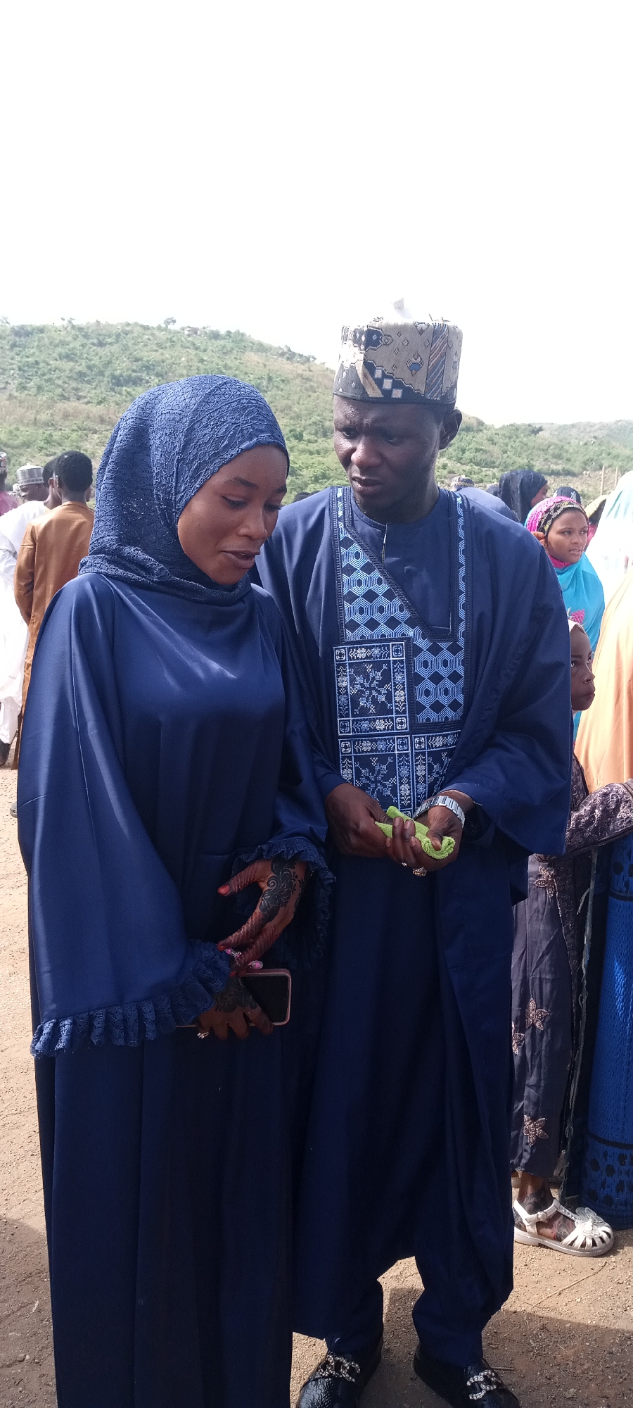 In Naraguta Hausawa, Nigeria, a man and a woman dressed in matching navy blue traditional attire stand together outdoors. The woman wears a hijab and holds a cell phone in her left hand, revealing henna designs on her hands. The man wears a patterned cap and a robe over a tunic with geometric designs. He holds a small green object in his hands. Other people are visible in the background. The environment appears to be a gathering or event with a slightly hilly landscape in the distance.