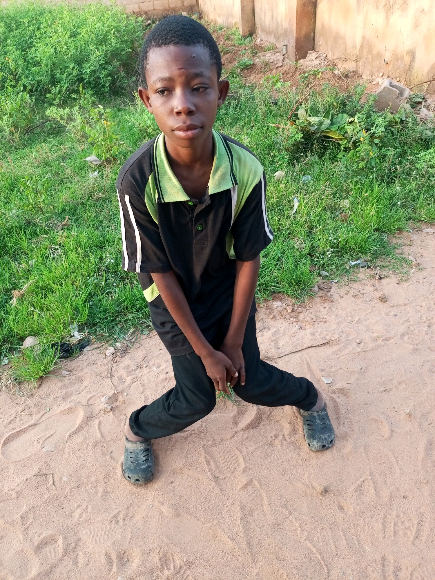 A young Black boy with dark skin and short black hair is standing in a sandy area in front of a brick wall and some vegetation. He is wearing a black polo shirt with light green accents on the collar and sleeves, and black pants. His hands are clasped together in front of him. He is wearing dark colored crocs. The ground is sandy with some footprints visible. The background shows a textured light-colored wall and green grass with some plants. The image appears to be taken outdoors during the day. The location is identified as Tempure, Nigeria.
