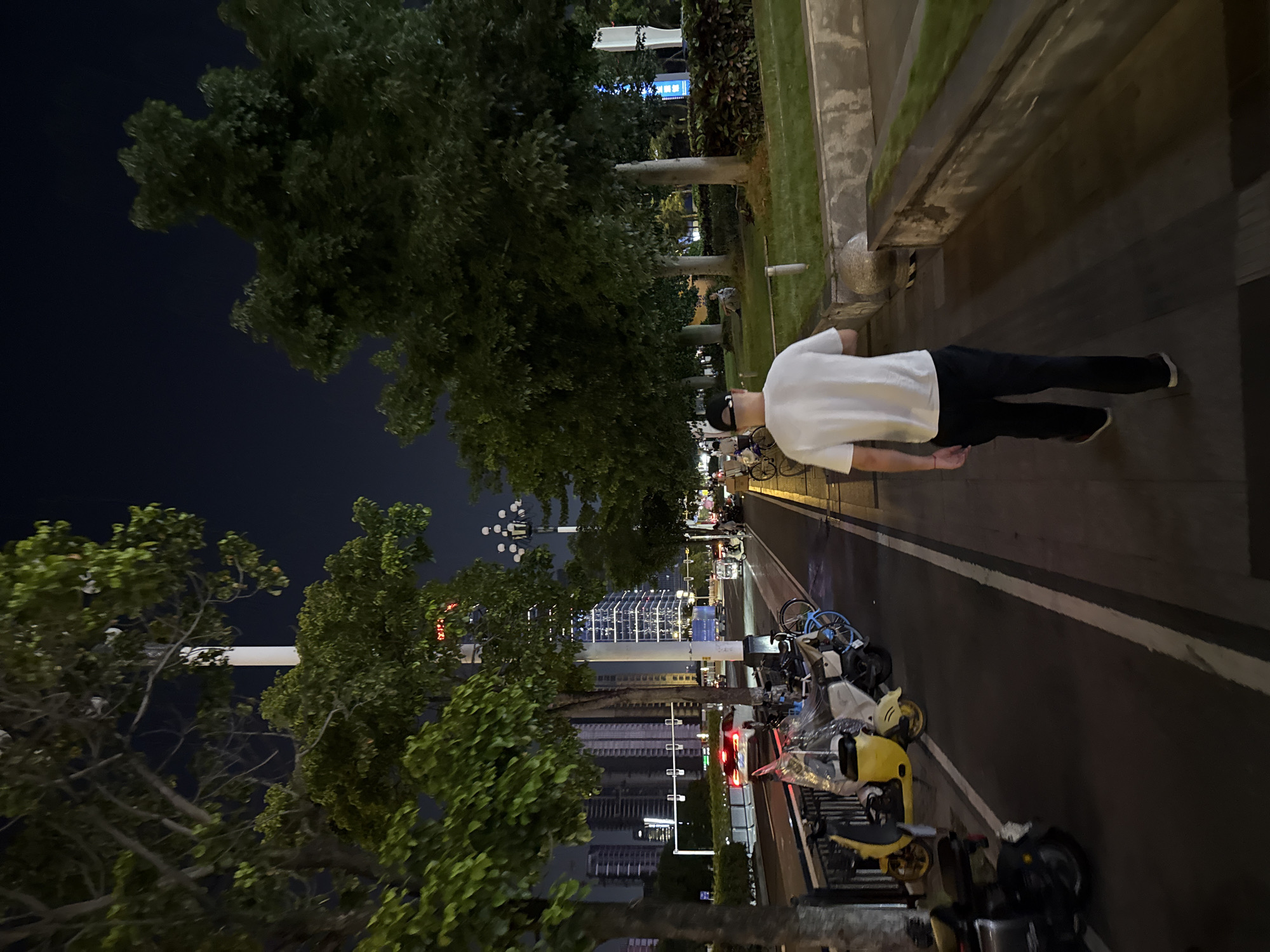 A man wearing a white t-shirt and black pants walks away from the camera on a paved sidewalk at night. He has a black baseball cap on. The sidewalk runs alongside a street with traffic, including cars and motorcycles. There are trees lining the sidewalk and street, with city buildings in the background. The image appears to be taken in Tianhe District, China, judging by the urban landscape and architectural style.