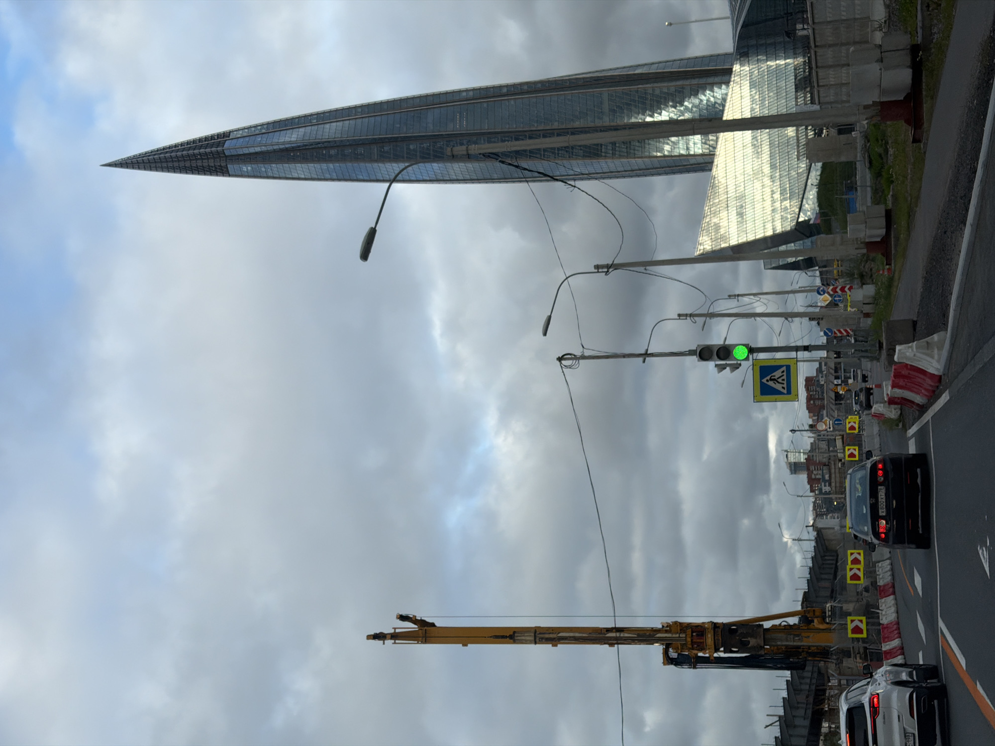 The image captures a view of the Lakhta Center skyscraper in Saint Petersburg, Russia, on an overcast day. The skyscraper, a prominent landmark, stands tall with its distinctive spire. In the foreground, a construction site is visible, featuring a large yellow drilling rig and temporary barriers. A traffic light is illuminated green, and a pedestrian crossing sign is posted. Several cars are present on the road, with one white SUV and a black sedan clearly visible. Power lines and streetlights are also noticeable, contributing to the urban scene. The sky is filled with heavy, gray clouds, indicating a potentially cloudy or rainy weather condition.