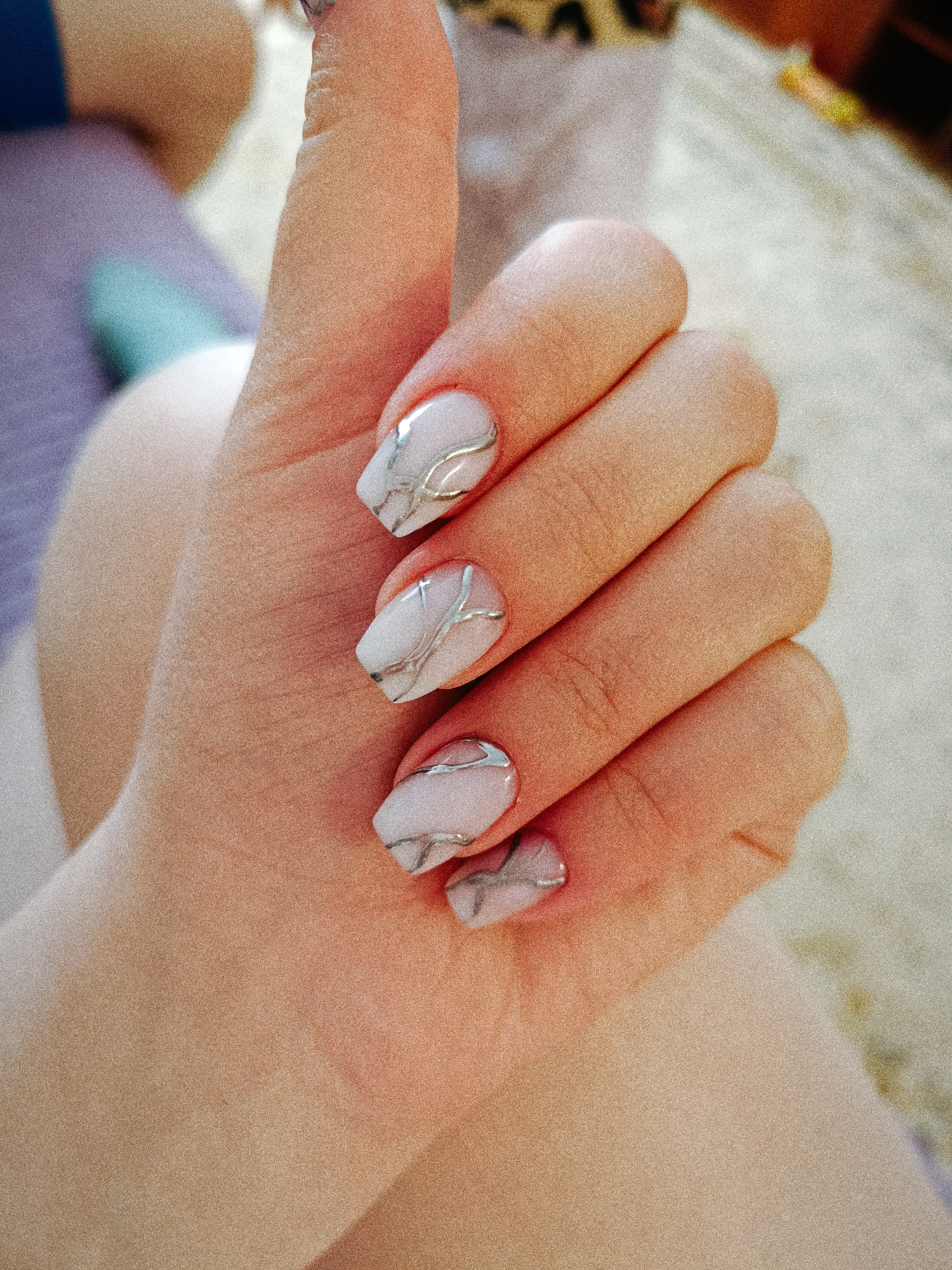 A close-up shot showcases a hand with a manicure, featuring a white base and silver veining on the nails. The hand is posed with the thumb extended upwards in a thumbs-up gesture. The background appears to be blurred, suggesting an indoor setting. There's a glimpse of a purple surface and a beige carpet. The image does not provide any information to suggest it was taken in Tobolsk, Russia.