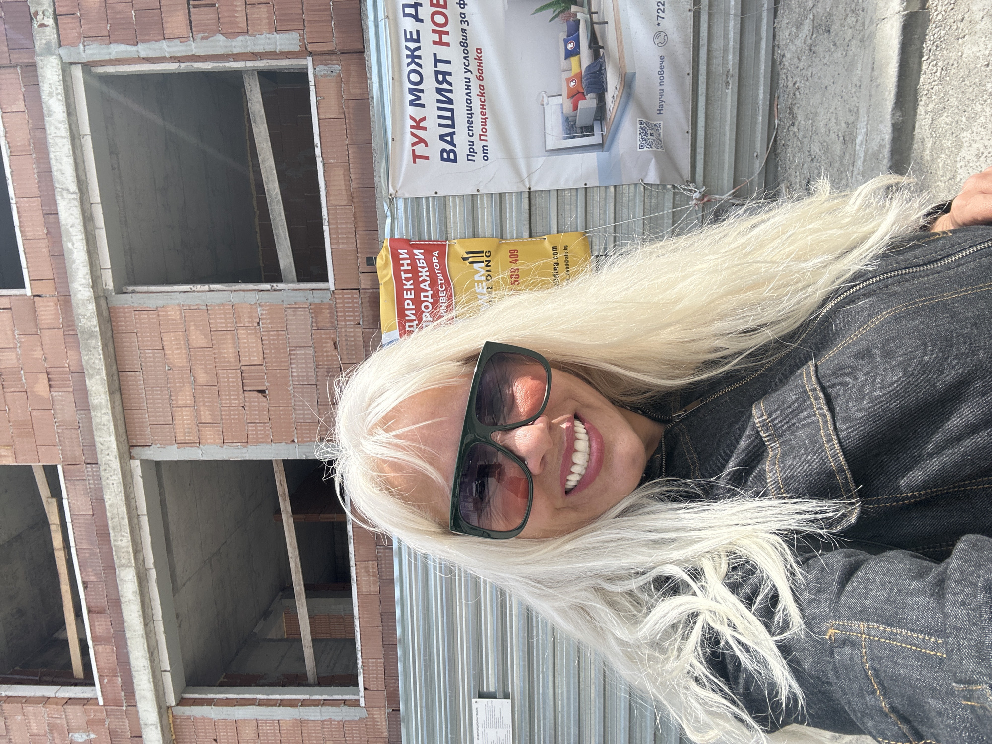 A woman with long blonde hair is smiling at the camera. She is wearing sunglasses and a denim jacket. Behind her is a brick building under construction with window openings. A corrugated metal fence is partially visible, and two signs are attached to it. One sign advertises "DIRECT SALES FROM THE INVESTOR" in Bulgarian, while the other, larger sign reads "HERE YOU CAN FIND YOUR NEW HOME" and mentions special conditions from Postbank. The sign also includes contact information and a QR code. The location is likely in Sofia, Bulgaria, due to the Bulgarian language on the signs.