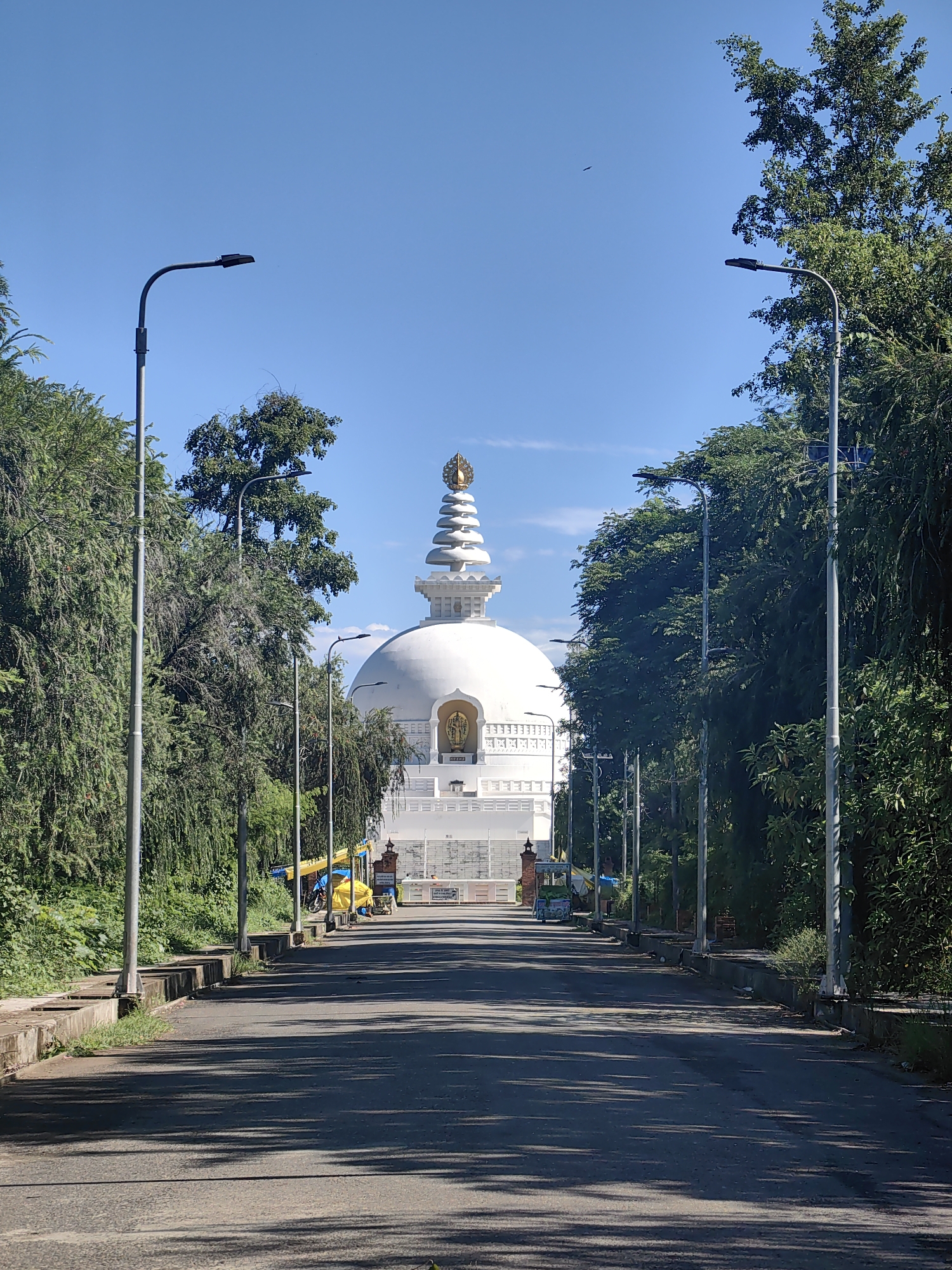 The image shows the World Peace Pagoda in Lumbini, Parsa, Nepal. It is a large, white stupa with a golden spire, surrounded by lush green trees. The stupa is situated on a hilltop, overlooking a valley. In the foreground, there is a road leading up to the stupa, with streetlights and a few parked vehicles. The sky is clear and blue, with a few clouds visible. The overall scene is serene and peaceful, indicative of a sacred site.