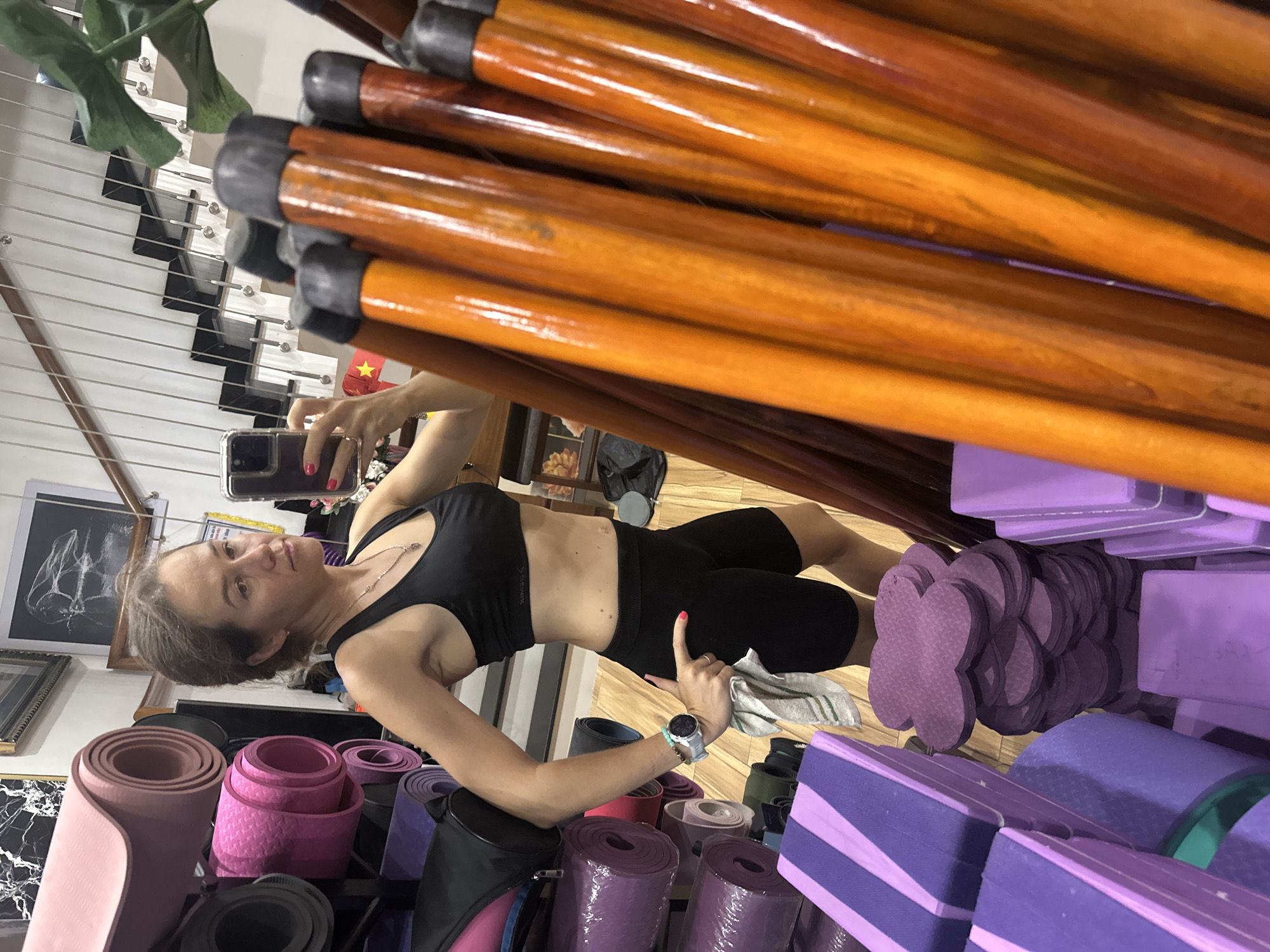 A woman is taking a selfie in what appears to be a yoga studio in Bac Nha Trang Ward, Vietnam. She is wearing a black sports bra and black shorts, holding a smartphone in her left hand. Her right hand is raised with a finger pointing. She has a watch on her left wrist.
The environment includes yoga mats of various colors (pink, purple, black) and yoga blocks in purple. There are wooden bars stacked in the background and to the right. A set of stairs is visible in the background on the left. A Vietnamese flag is visible on the staircase. Framed pictures hang on the wall.