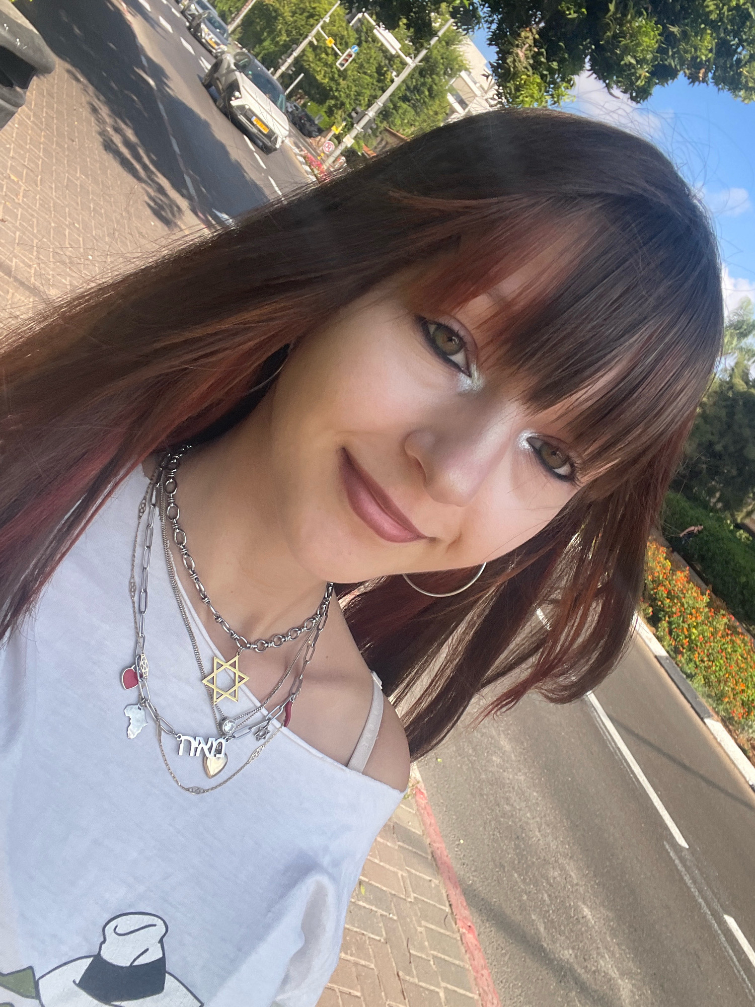 A young woman with long, dark brown hair and bangs is taking a selfie outdoors. She is wearing a white off-the-shoulder top adorned with multiple silver necklaces, including one with a Star of David pendant and another with Hebrew lettering. Her makeup includes silver eyeshadow and lip gloss. The background suggests an urban environment in Raanana, Israel, with a paved sidewalk, a road with cars, trees, and a flowerbed with orange and red flowers. The sky is blue with some clouds. The overall scene is a casual, daytime snapshot.