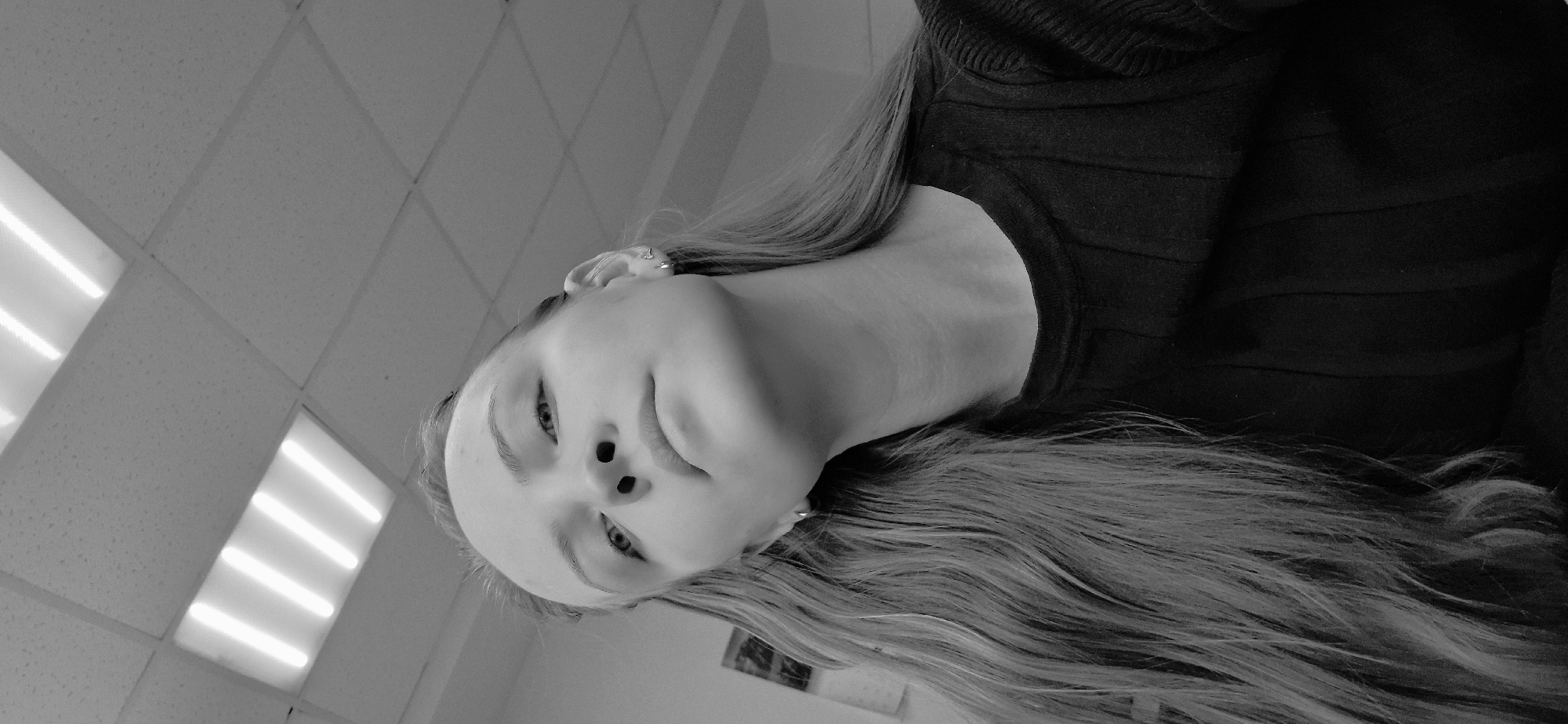 The monochromatic image presents a female individual positioned in an indoor environment. She is captured from a low-angle perspective, with her head tilted slightly backward, and her gaze directed upwards and to the left. Her hair is long, light-colored, and flows down her shoulders and chest. She is wearing small earrings in both ears and a dark, vertically ribbed long-sleeved top. In the background, a light-colored wall is partially visible, displaying a framed item, possibly a picture or calendar, on the left side of the frame. The ceiling above features a grid of acoustic tiles, housing multiple illuminated recessed fluorescent light fixtures, each containing several parallel light tubes. The specific location within the city of Chaikovsky, Russia, cannot be determined from the visual information provided.