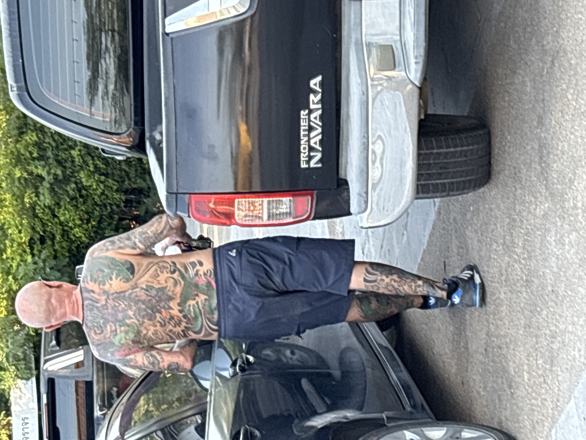 The image depicts a man with a bald head and extensive tattoos covering his back and arms standing next to the rear of a black "FRONTIER NAVARA" pickup truck. He is wearing dark blue shorts and blue and black sneakers. The location appears to be a street in บ้านบางรักษ์, Thailand, given the presence of Thai writing on a sign at the lower left of the frame. The man is interacting with a black car parked adjacent to the truck. The environment includes a paved road, a vehicle, and foliage.