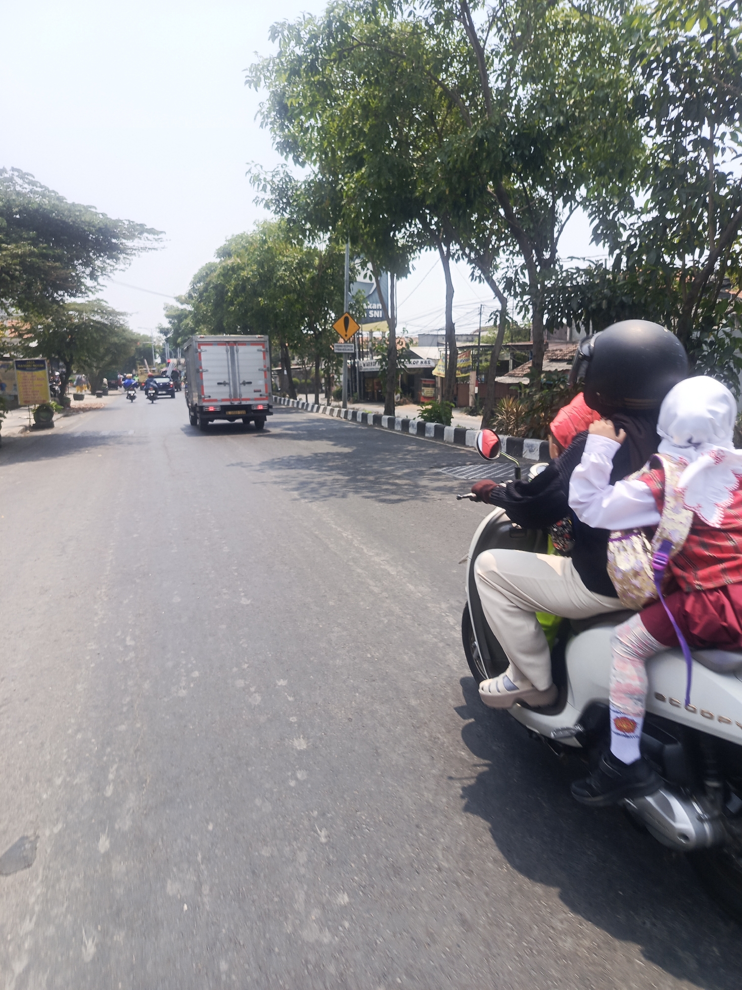 The image captures a street scene in Surabaya, Indonesia, on a sunny day. In the foreground, two individuals are riding a white scooter. The person in the back, a child, wears a red and white outfit, a white hijab, and patterned socks. They are holding onto the driver, who is wearing a black helmet, a red headscarf, a black top, and light-colored pants. The scooter is moving along an asphalt road with some debris and water stains.
In the mid-ground, a large white truck is driving away from the camera. Further down the road, other vehicles and motorbikes are visible, as well as pedestrians on the sidewalk. The environment features trees lining the street, some buildings, and a bright sky. A yellow traffic sign with a black exclamation mark is visible on the right side of the road. The overall impression is a typical urban street with moderate traffic under a clear sky.