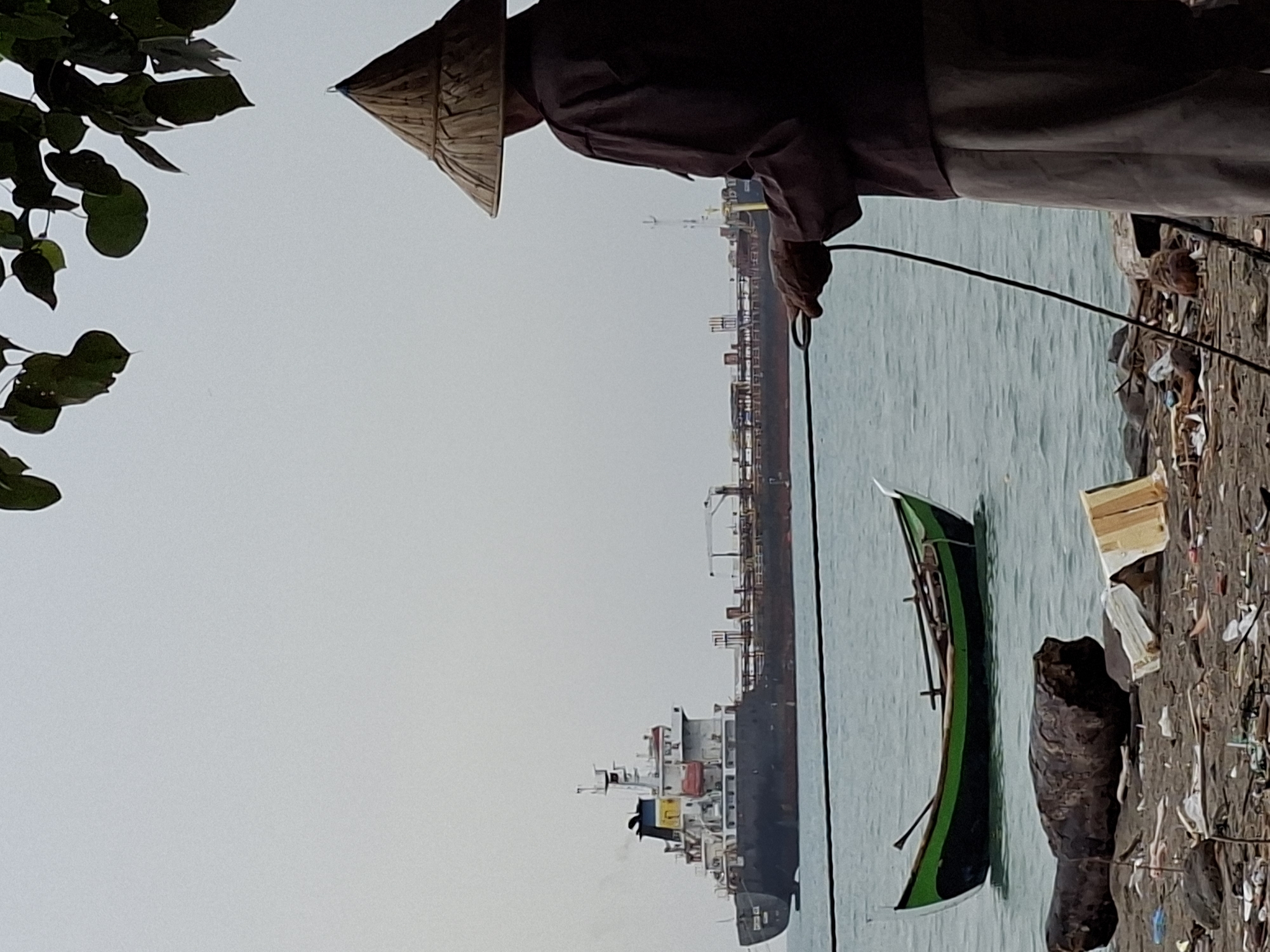 An image taken in Lhokseumawe, Indonesia, shows a person wearing a conical hat and dark clothing on a beach. The person is holding a rope, with their back to the camera. In the foreground, a green and black boat is floating on the water. The beach is littered with debris, including logs and trash. In the background, a large cargo ship is anchored in the sea under an overcast sky.