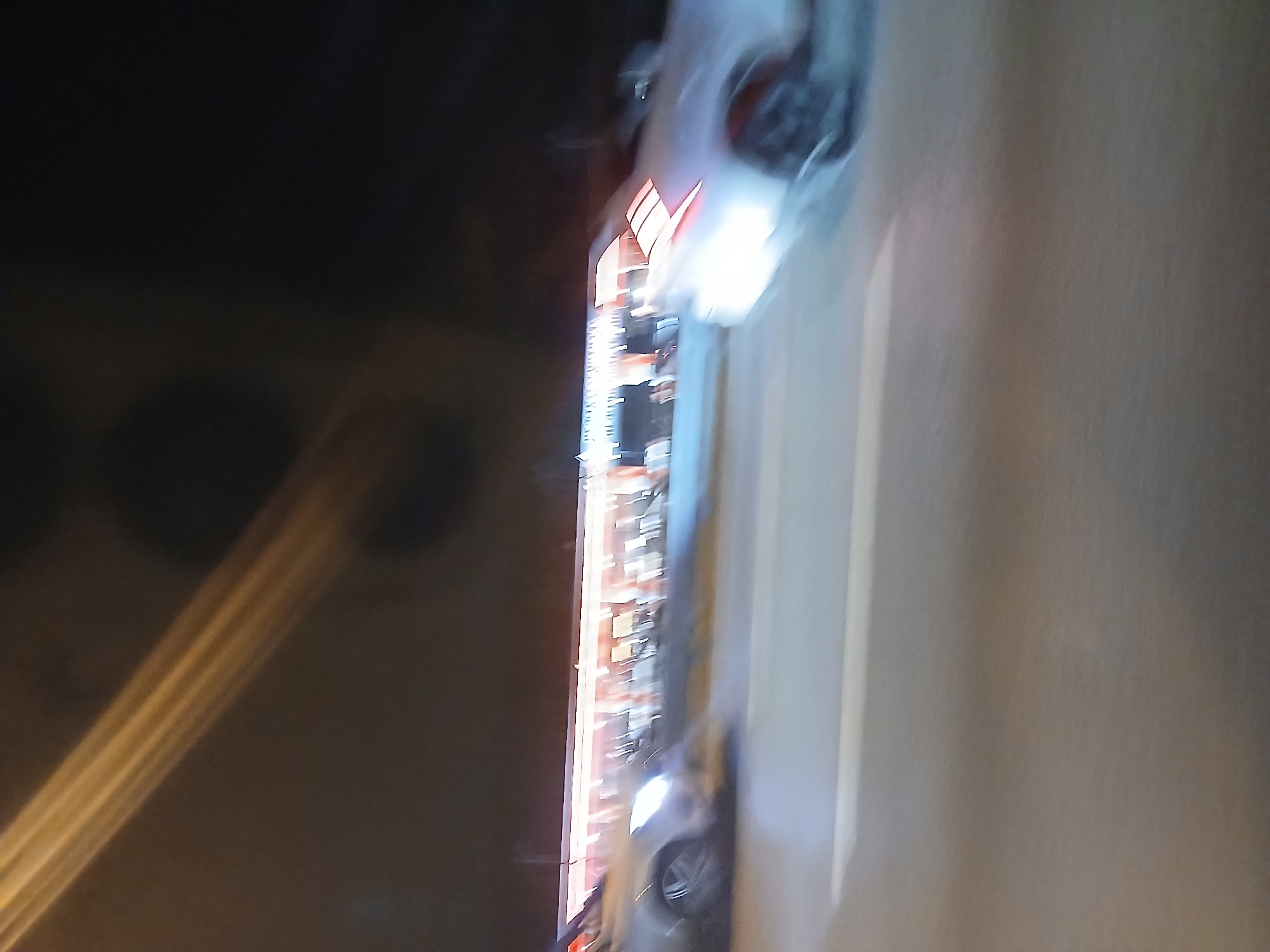 The image depicts a nighttime scene on a paved road in the city of Unknown, Unknown, exhibiting significant motion blur throughout. The foreground consists of a dark gray asphalt surface with a faint white lane marking visible horizontally in the lower center.
In the mid-ground, a commercial building or establishment is visible, featuring a brightly illuminated facade with numerous horizontally arranged light sources, appearing as a continuous band of light. Below this illuminated section, an indistinct outdoor seating area with tables and chairs is suggested.
Two light-colored vehicles, consistent with sedans or coupes, are depicted in motion on the road. A vehicle is positioned in the left mid-ground, with its illuminated front headlight visible, along with a multi-spoke wheel. A second vehicle occupies the right mid-ground, displaying illuminated rear taillights. Both vehicles show motion blur indicative of forward movement.
The upper portion of the frame shows a dark night sky. A prominent golden-orange diagonal light streak extends from the upper left towards the center. A dark, roughly circular aberration is visible in the upper-center frame. No discernible individuals are visible. Illumination sources include vehicle lights and the lights from the distant building.