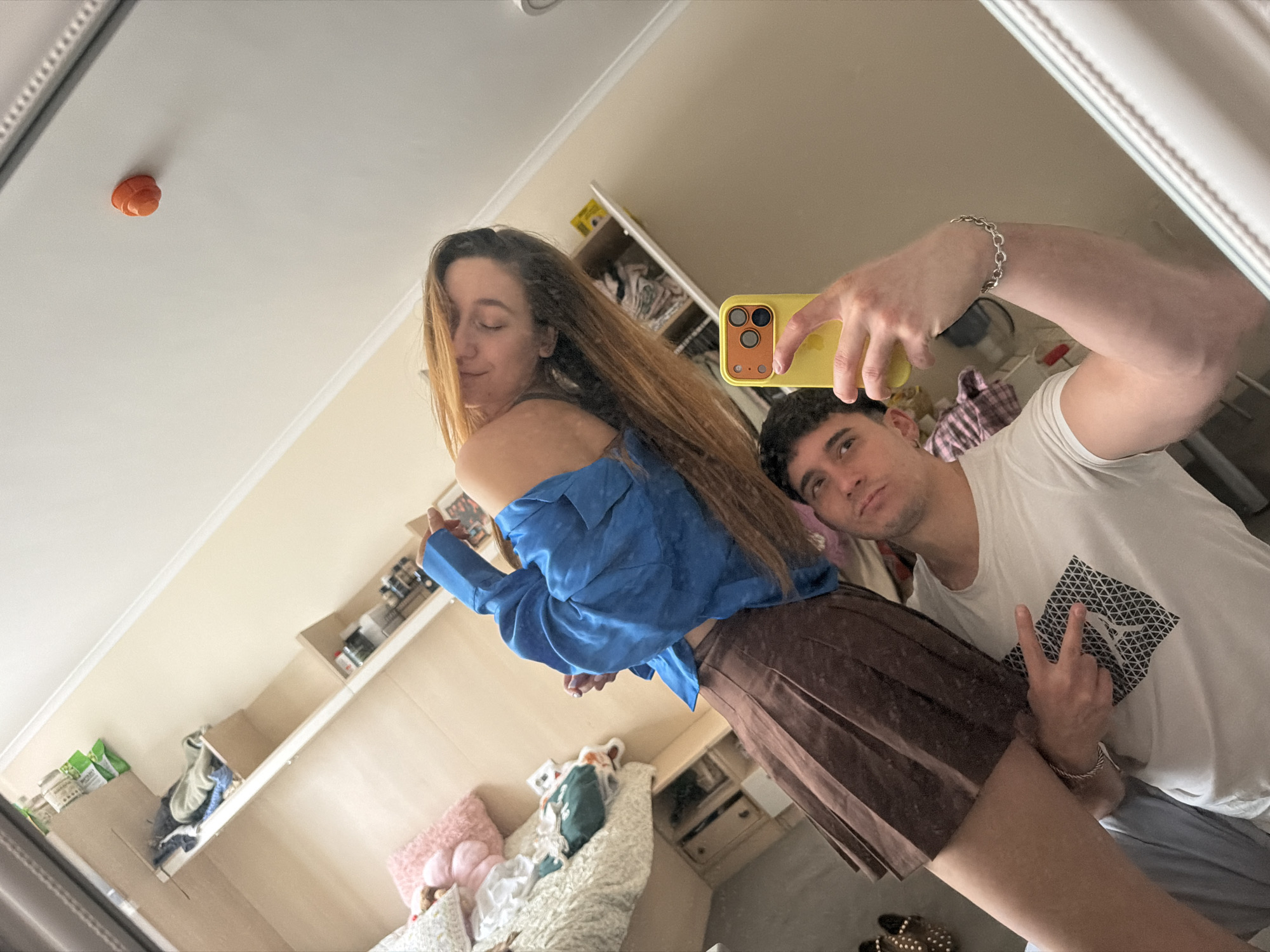 A man and a woman are taking a selfie in front of a mirror. The woman has long brown hair, wearing a blue top and a brown skirt. The man is holding a yellow phone, wearing a white t-shirt, and making a peace sign with his hand. The background includes shelves with various items, a bed with pillows, and an orange object on the ceiling. There is no specific information to identify the location as Sofia, Bulgaria.