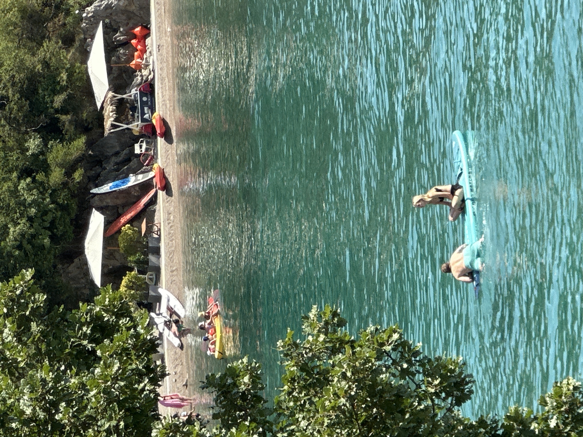 The image depicts a scenic view of a beach and clear turquoise water in Budva, Montenegro. In the foreground, two individuals are on a paddleboard in the water. A man is lying on his stomach on the front of the paddleboard, and a woman is sitting cross-legged on the back.
In the mid-ground, a yellow kayak with several people in it is moving across the water. Further back, a sandy beach is visible with several lounge chairs, umbrellas, and water sports equipment, including surfboards and kayaks. A small bar or service stand with a thatched roof is also present on the beach. People are seen walking and relaxing on the sand. Lush green trees and rocky cliffs form the backdrop of the beach. The water is calm and reflects the sunlight, creating a sparkling effect.