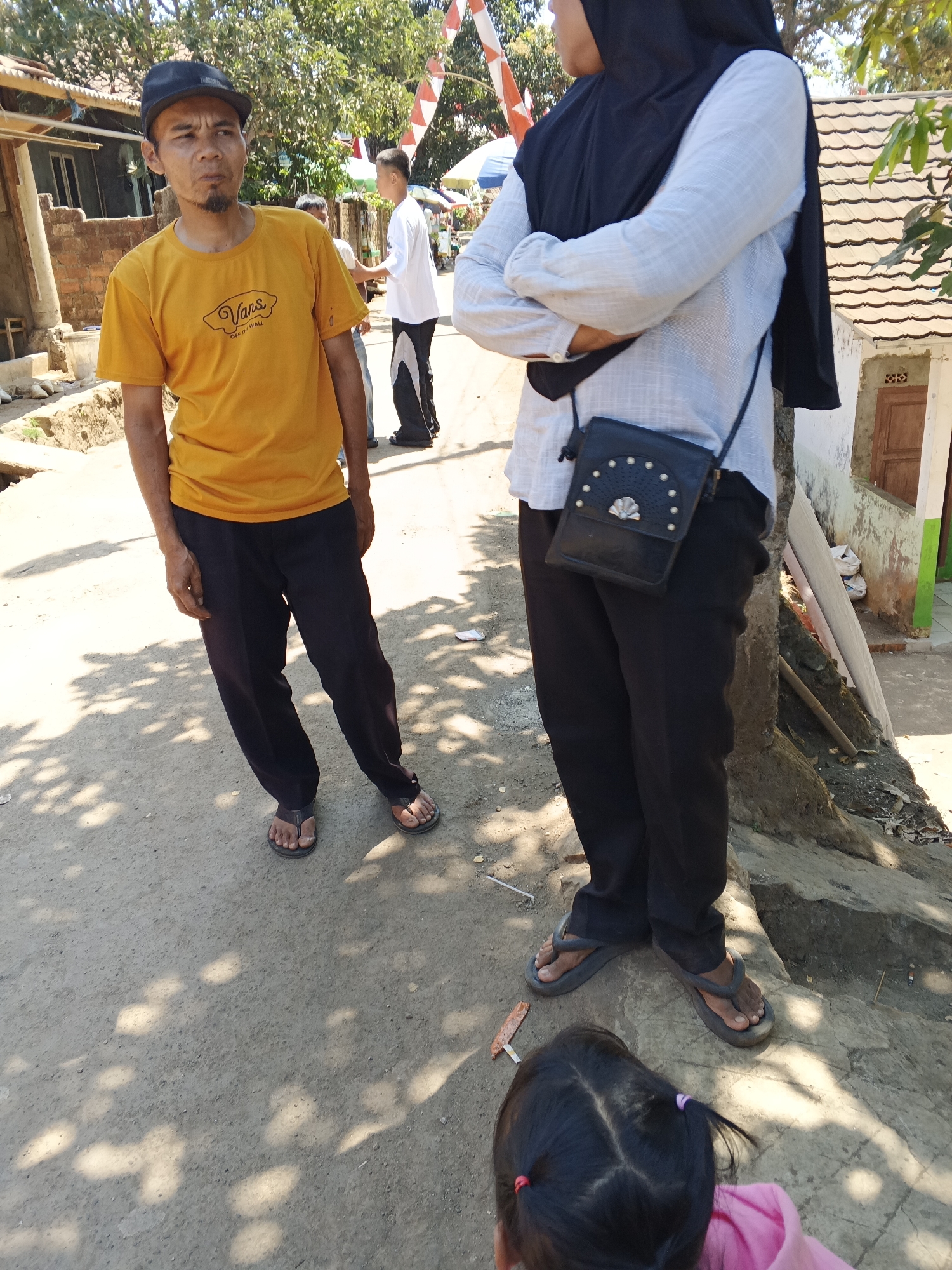 In Sukaharja, Indonesia, on a sun-drenched dirt road, three individuals are captured in a candid moment. A man in his late 30s, sporting a black baseball cap and a yellow t-shirt with a "Vans" logo, stands with his hands in his pockets, his gaze directed slightly away from the camera, a subtle goatee adorning his chin. To his right, a woman, her face obscured by a black hijab, is seen from the waist up, her arms crossed and a small black purse with decorative studs slung across her chest. Her light blue, long-sleeved shirt is casually worn. In the background, another man in a white t-shirt and dark pants is partially visible. The environment suggests a rural or semi-urban setting with a simple brick building on the left and a tiled-roof structure on the right, adorned with festive red and white banners. The ground is a mixture of dirt and scattered leaves, with dappled sunlight casting shadows from overhead foliage. A young child, facing away from the viewer and wearing a pink top, is also present, her dark hair tied with purple and red ribbons.