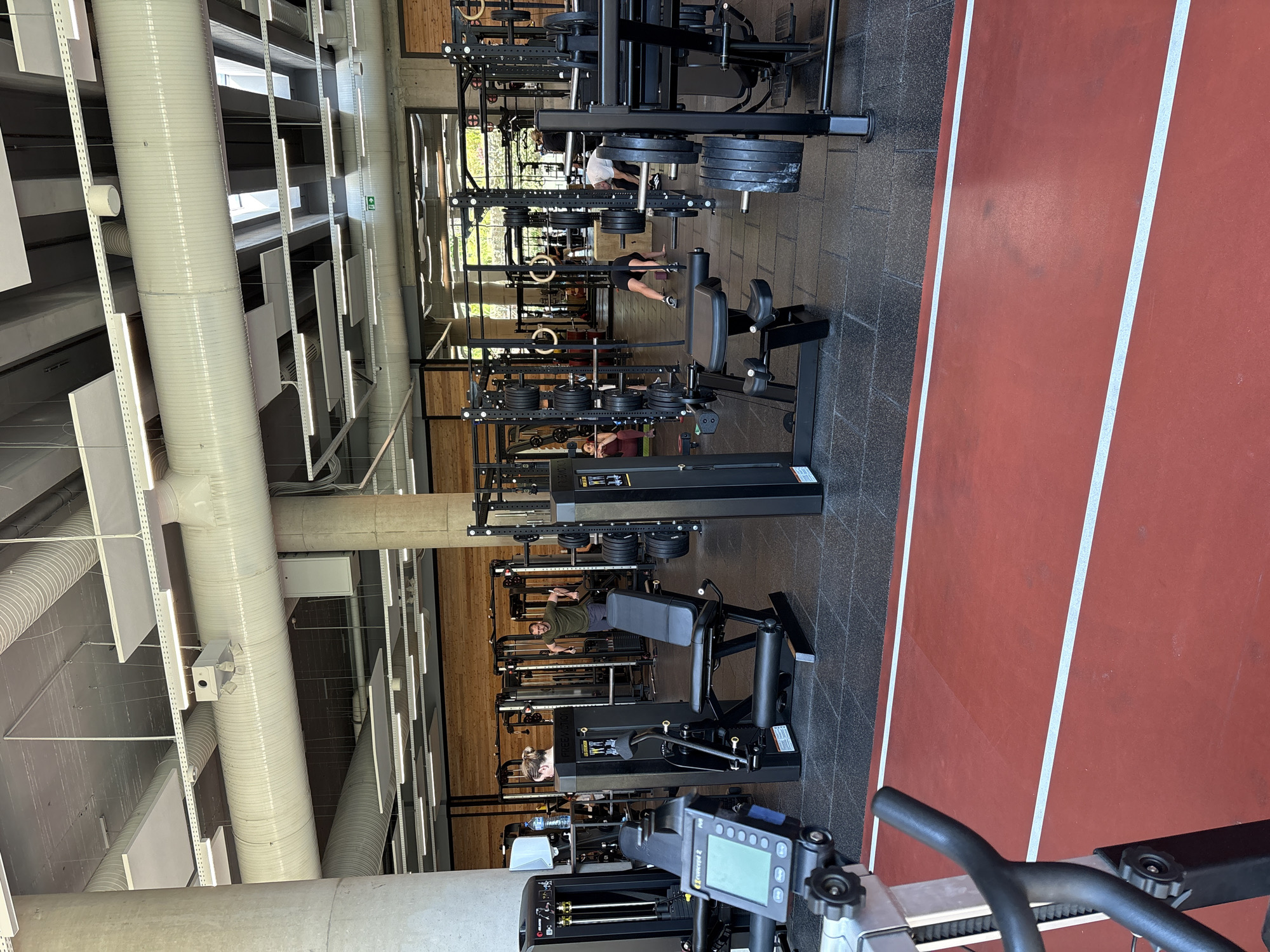 This image depicts the interior of a modern gym or fitness center in Sofia, Bulgaria, during what appears to be daytime, indicated by natural light streaming through windows. The scene is viewed from a slightly elevated perspective, looking across the gym floor.
The space features an industrial-chic design, with a high ceiling displaying exposed large white ventilation ducts, concrete beams, and suspended white sound-absorbing panels. Concrete pillars are also visible, complementing the wooden paneling on some walls.
Several individuals are present, actively engaged in various workout activities. One person is using a leg press machine, another is seen performing floor exercises, and others are near weight racks with various weight plates. The gym is well-equipped with numerous black weight machines, free weights, and barbells. In the foreground, a black and red rowing machine, identified by the visible "Concept2" branding and its digital display, is positioned ready for use.
The flooring consists of dark gray rubber matting in the primary workout areas, transitioning to a distinct red athletic track marked with two parallel white lines on the right side of the frame. Visible text includes "Concept2" on the rowing machine and "FREEMOTION" on some of the weight machines. A green directional arrow sign is also discernible on a distant wall. The overall ambiance is one of active physical training.