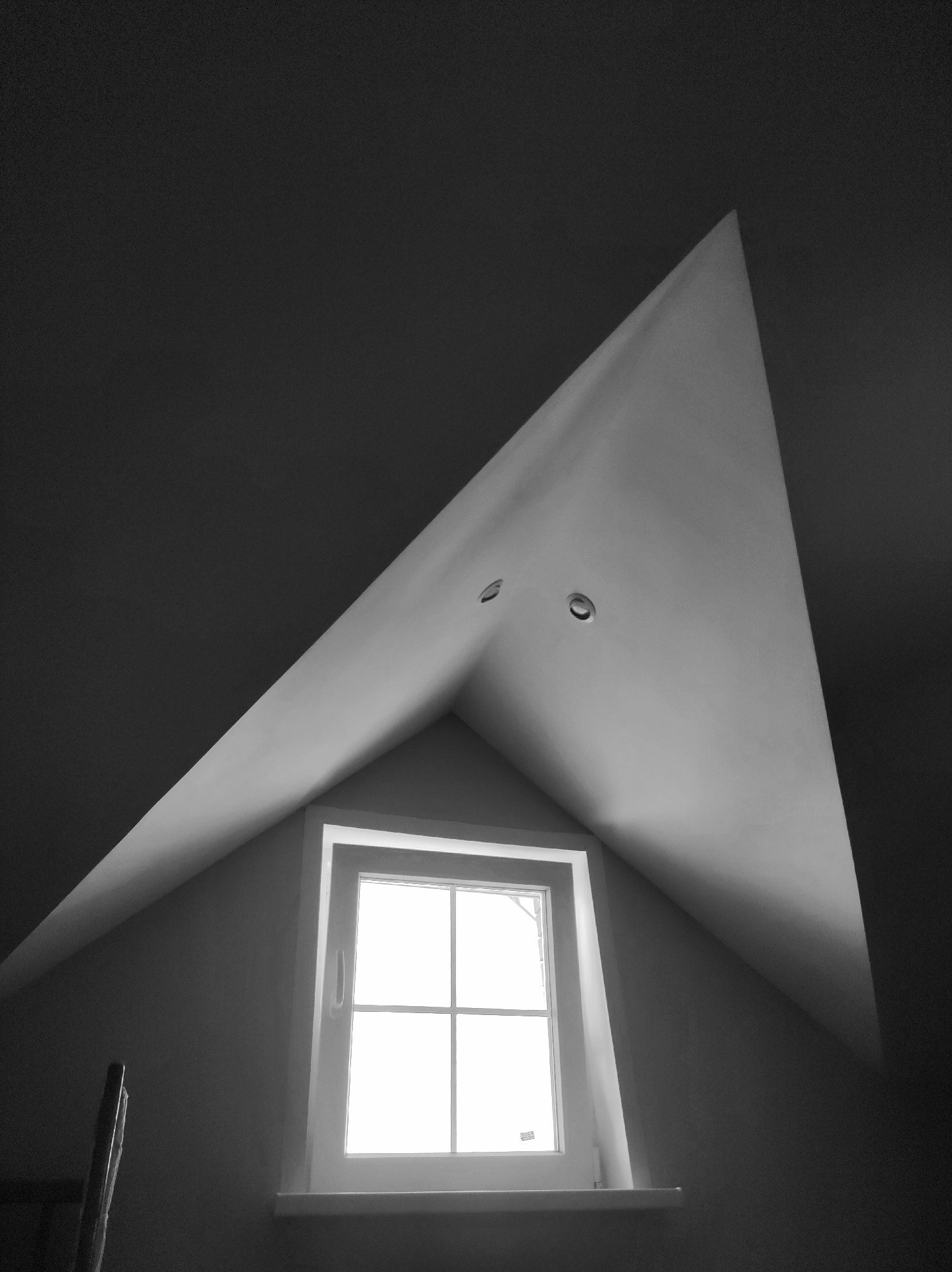 A monochrome, low-angle shot depicts a room with a triangular dormer window. The window is bright, suggesting daylight, and is centered within the triangular space created by the sloping roof. Two recessed lights are visible in the ceiling above the window. The environment appears to be an interior space, possibly an attic or upper floor room, with dark walls contrasting with the lighter tones of the dormer and window frame. A portion of what appears to be a ladder is visible in the bottom left corner. No people or specific actions are depicted. Location: Unknown, Unknown.