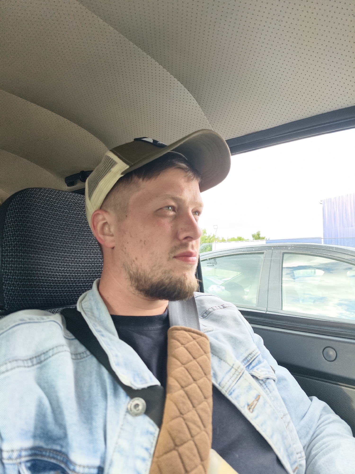 A man sits in a car, wearing a denim jacket, a dark t-shirt, and an olive green baseball cap. He has a short haircut and a beard. His seatbelt is fastened and has a brown quilted padding on it. The car's interior is gray, and the car is parked. Through the car window, a silver car and a large blue building are visible. The location is unidentified.