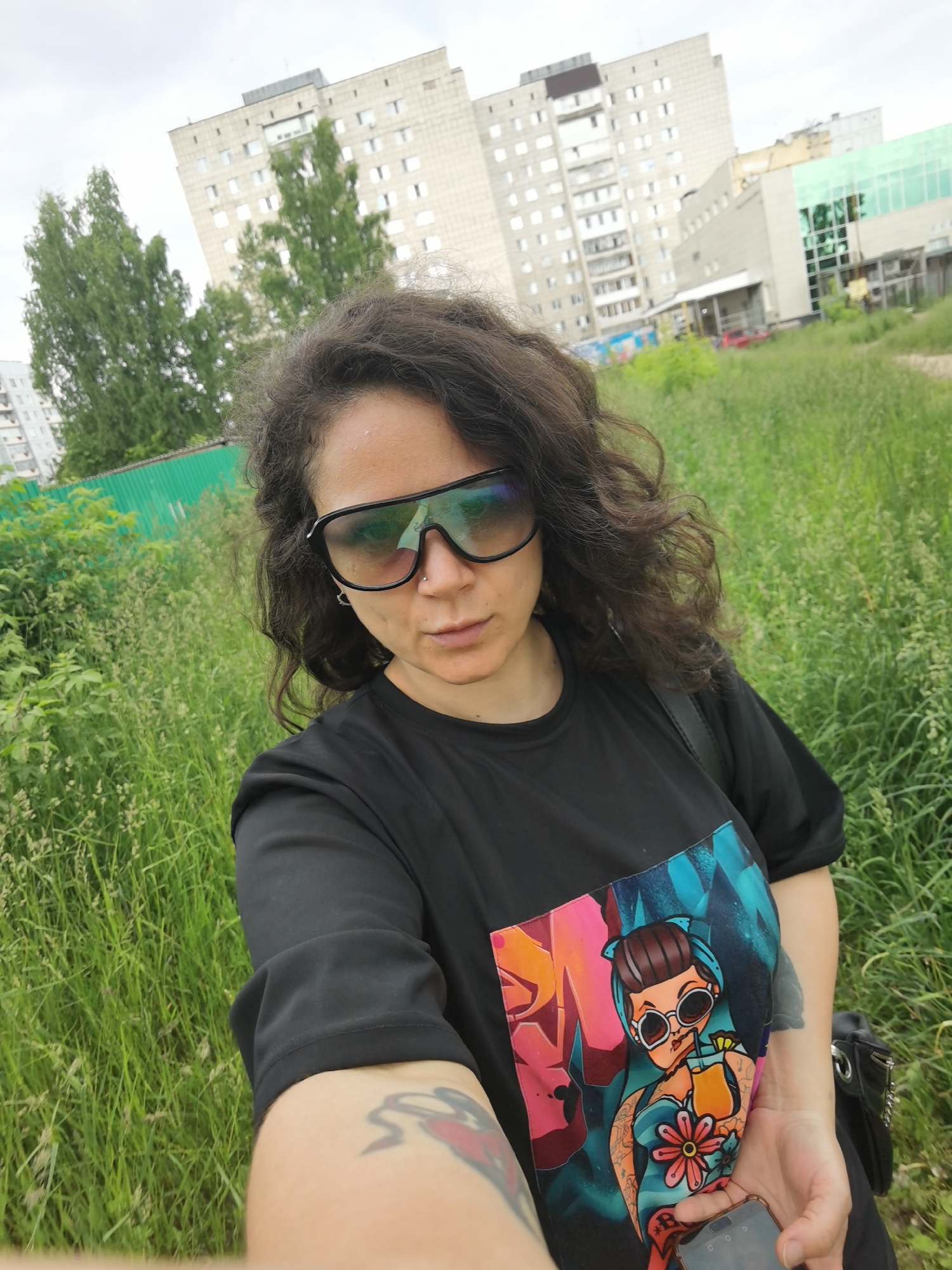 A woman with dark, curly hair and sunglasses is taking a selfie outdoors in Chaikovsky, Russia. She is wearing a black t-shirt with a colorful graphic of a cartoon character. The background shows tall buildings and lush green grass. The sky is overcast.