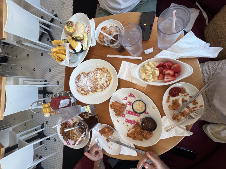 A round wooden table sits in a cafe in Miami Beach, Florida, at the coordinates 25.771, -80.134. Three people are seated around the table, and there are several plates of food, glasses, and utensils on the table. One person is holding a fork and knife. Two plates of food are visible in the foreground, containing fried chicken and french toast. A third plate, partially visible, contains a variety of toppings, including syrup, whipped cream, and powdered sugar. Another plate, partially visible, contains a large burrito with a side of chips and salsa. Three glasses of water and a glass of iced coffee are also visible on the table. There are also several napkins, condiments, and paper place mats on the table. The chairs are white and the floor is tiled. The cafe is brightly lit and the atmosphere is casual.