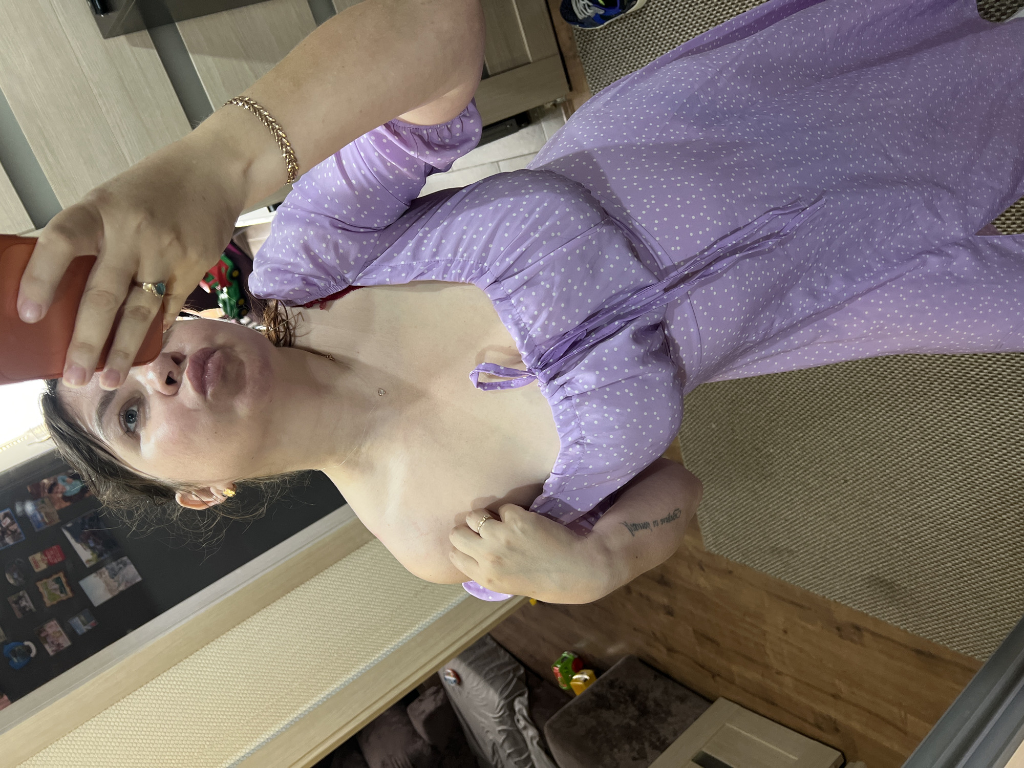 A woman is taking a mirror selfie in what appears to be an indoor setting, possibly a bedroom or living room, in Noviy, Russia. She is wearing a light purple, polka-dotted dress with a sweetheart neckline and tie detail. She is holding a red phone in her right hand, taking the picture. Her left hand is resting on her chest, and she is looking at her reflection with her lips pursed. She is wearing a ring on her left ring finger and a bracelet on her right wrist. In the background, a wooden floor and a textured rug are visible. There is also a glimpse of furniture and a door in the background.