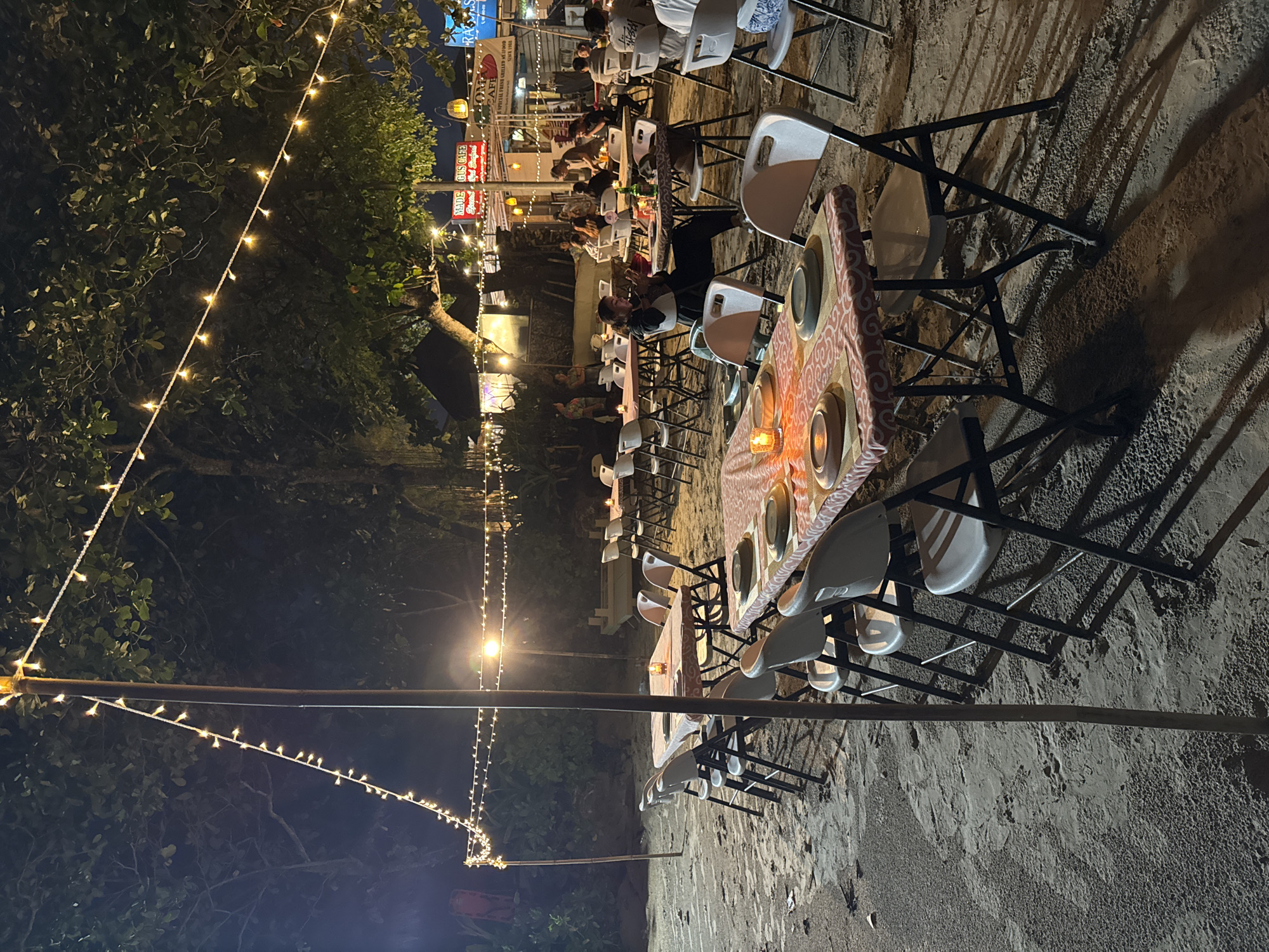 The image shows a nighttime scene in Kuta Selatan, Indonesia. Several long tables are set up on a sandy area, likely a beach or outdoor dining space, with white plastic chairs arranged around them. The tables are covered with patterned tablecloths and set with plates, indicating they are prepared for dining. People are seated at the tables, suggesting a restaurant or event. String lights are strung overhead, illuminating the scene. Trees are visible in the background, along with buildings and signs along the street. The sand around the tables shows the imprints of chairs and feet. A light pole with attached string lights runs along the left side of the image, further contributing to the ambiance. The overall impression is of an outdoor dining area with a festive atmosphere.
