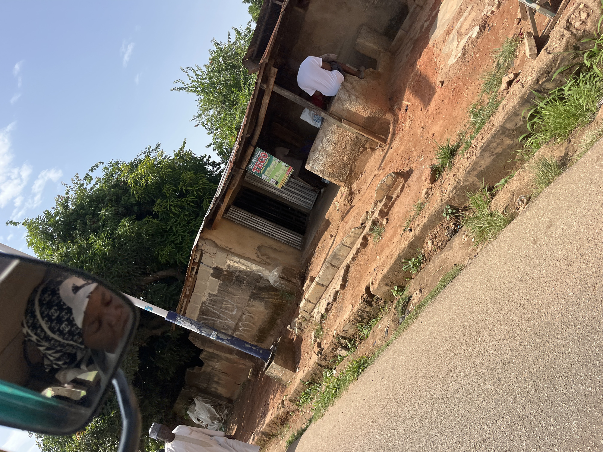 The image depicts a scene in Gura Top, Nigeria. In the foreground, a paved road runs diagonally from the bottom left to the upper left. To the right of the road, there is a dirt path with a shallow concrete drainage ditch. Beyond the path, a small, rustic building with a corrugated metal roof and a dirt floor is visible. The building has an open entrance and a sign advertising "Dr. Nwabu TECO HERBAL PRODUCTS." Inside, there are bags and shelves with products. A person in a white t-shirt is sitting on a raised concrete structure outside the building, facing away from the camera. Further left, a man wearing a white kaftan and a cap is walking towards the building. In the reflection of a rearview mirror in the upper left corner, a person wearing a headscarf is visible. The environment is characterized by red earth, sparse vegetation, and trees in the background under a bright, partly cloudy sky.