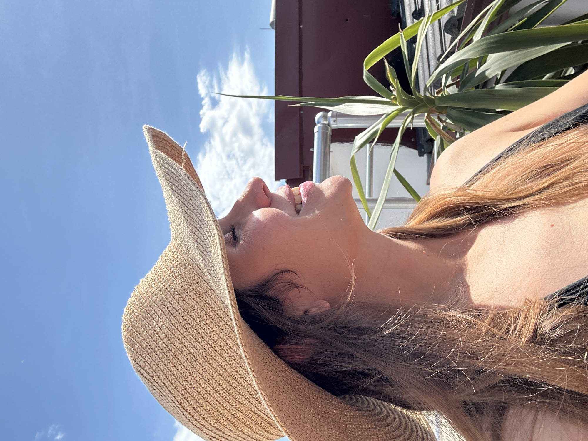 A low-angle, close-up shot looking upwards at a woman with fair skin and long, reddish-brown hair. She is wearing a wide-brimmed straw hat. The sky is visible, with scattered white clouds and a clear blue expanse. Behind the woman, a plant with long, pointed leaves can be seen. The background also includes a building with a dark red roof and a white wall with metal pipes. Based on the location, the photo was taken in Fethiye, Turkey.