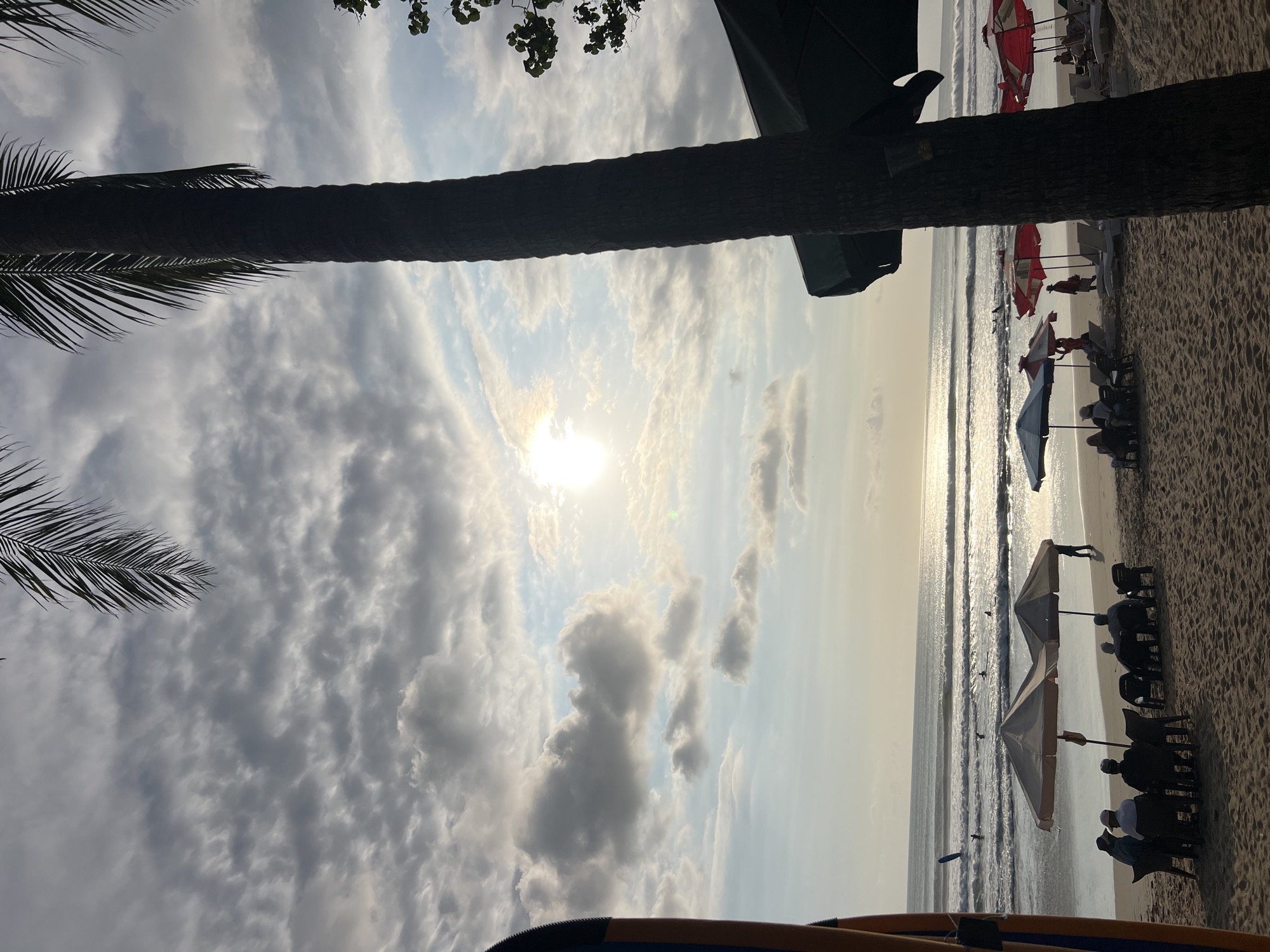 The image depicts a beach scene in Kuta, Indonesia, at sunset. The sky is filled with a mix of dramatic clouds and patches of blue, with the sun low on the horizon, casting a warm glow on the water. Palm trees frame the scene, with one tall palm trunk prominently in the foreground.
On the sandy beach, several groups of people are visible. In the middle ground, a cluster of chairs and umbrellas are set up, with individuals seated under them, facing the ocean. Further down the beach, more umbrellas and lounge chairs are arranged, with a few people relaxing. In the water, several surfers are scattered across the waves, enjoying the conditions. The ocean is calm with gentle waves rolling towards the shore. The overall atmosphere is peaceful and serene.